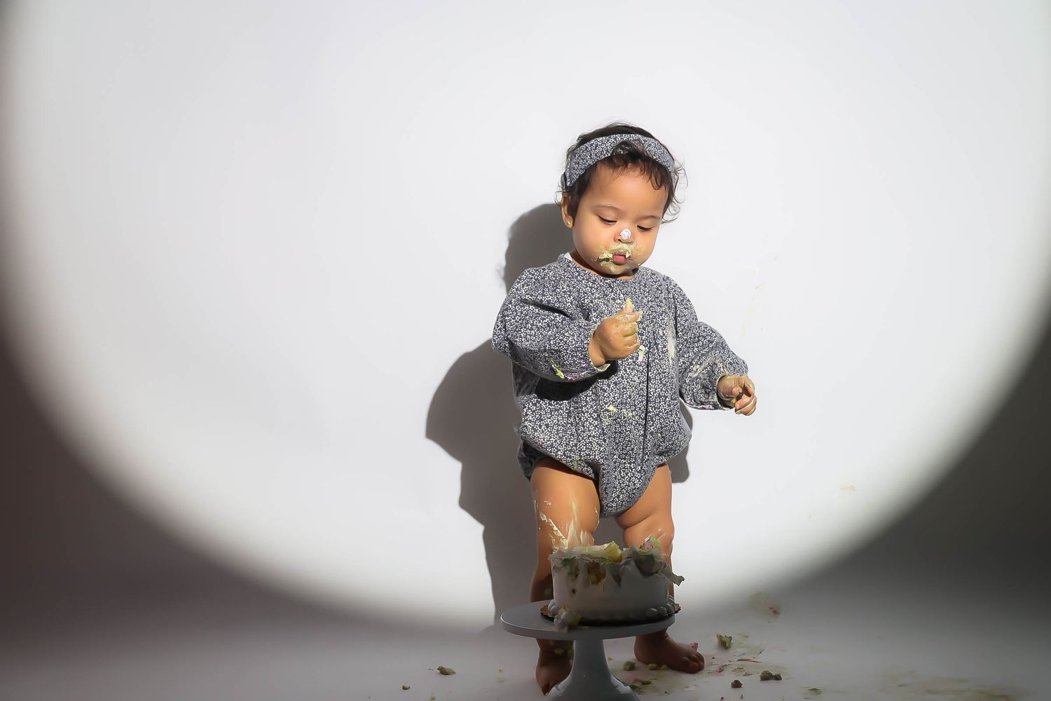 A baby in a gray knit outfit and bonnet stands holding a small bucket against a white background.