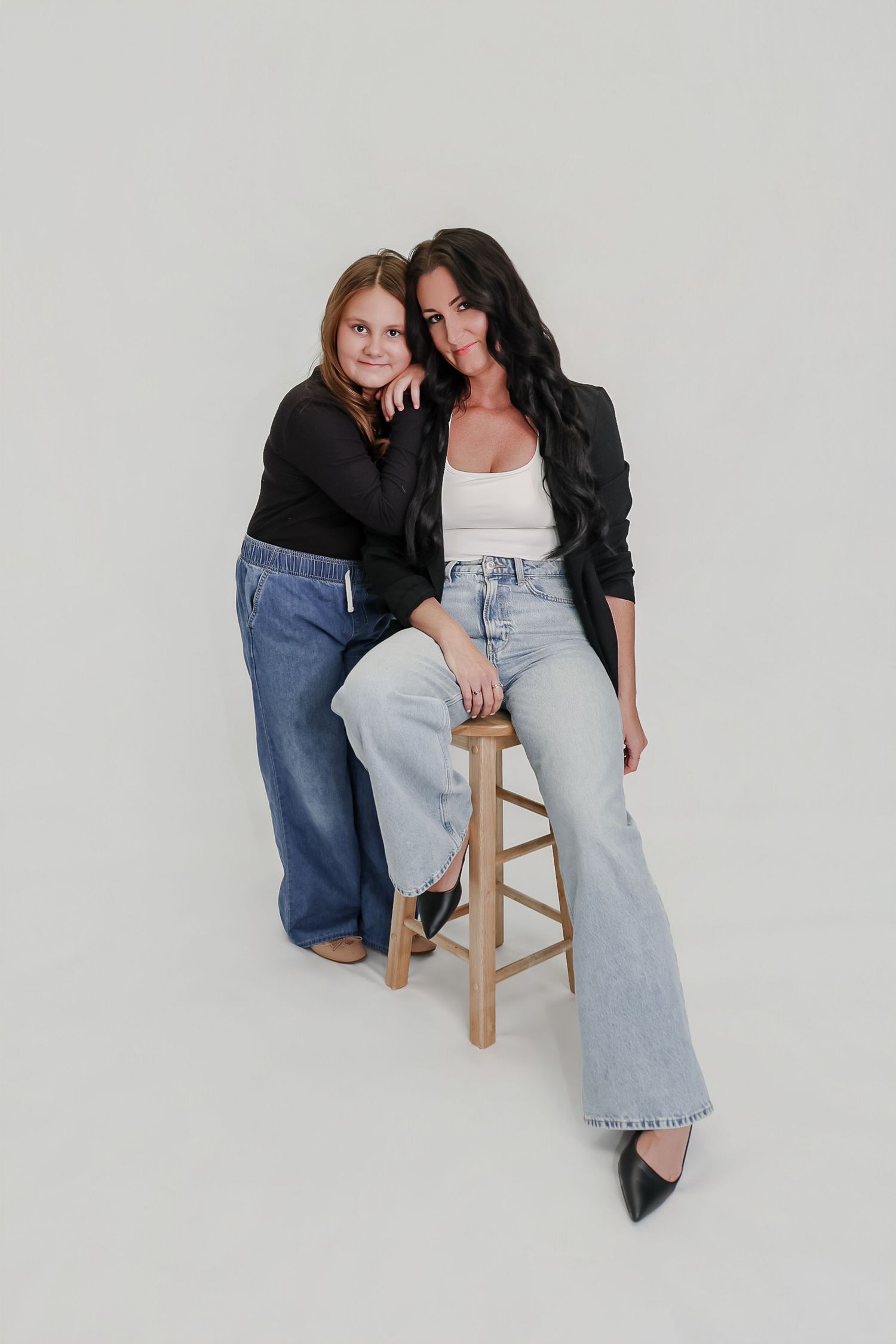 A woman in a black blazer sits on a stool beside a young girl, both wearing jeans, posing against a white background.