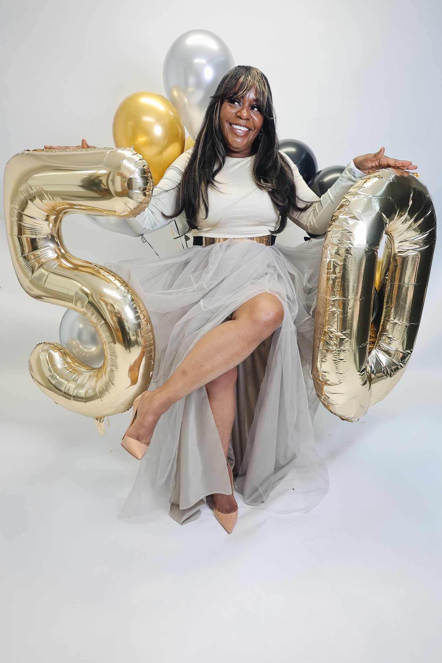 Woman celebrating 50th birthday with gold number balloons, wearing white top and gray tulle skirt, smiling joyfully.