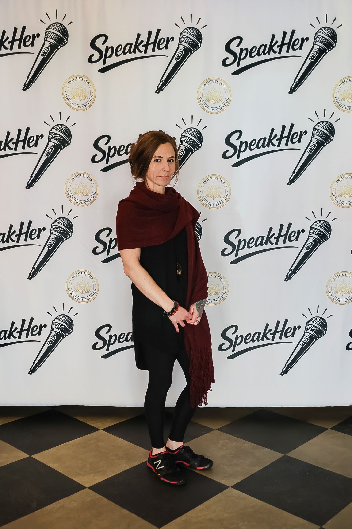 Woman in black outfit with burgundy shawl posing at SpeakHer event backdrop with microphone logos.