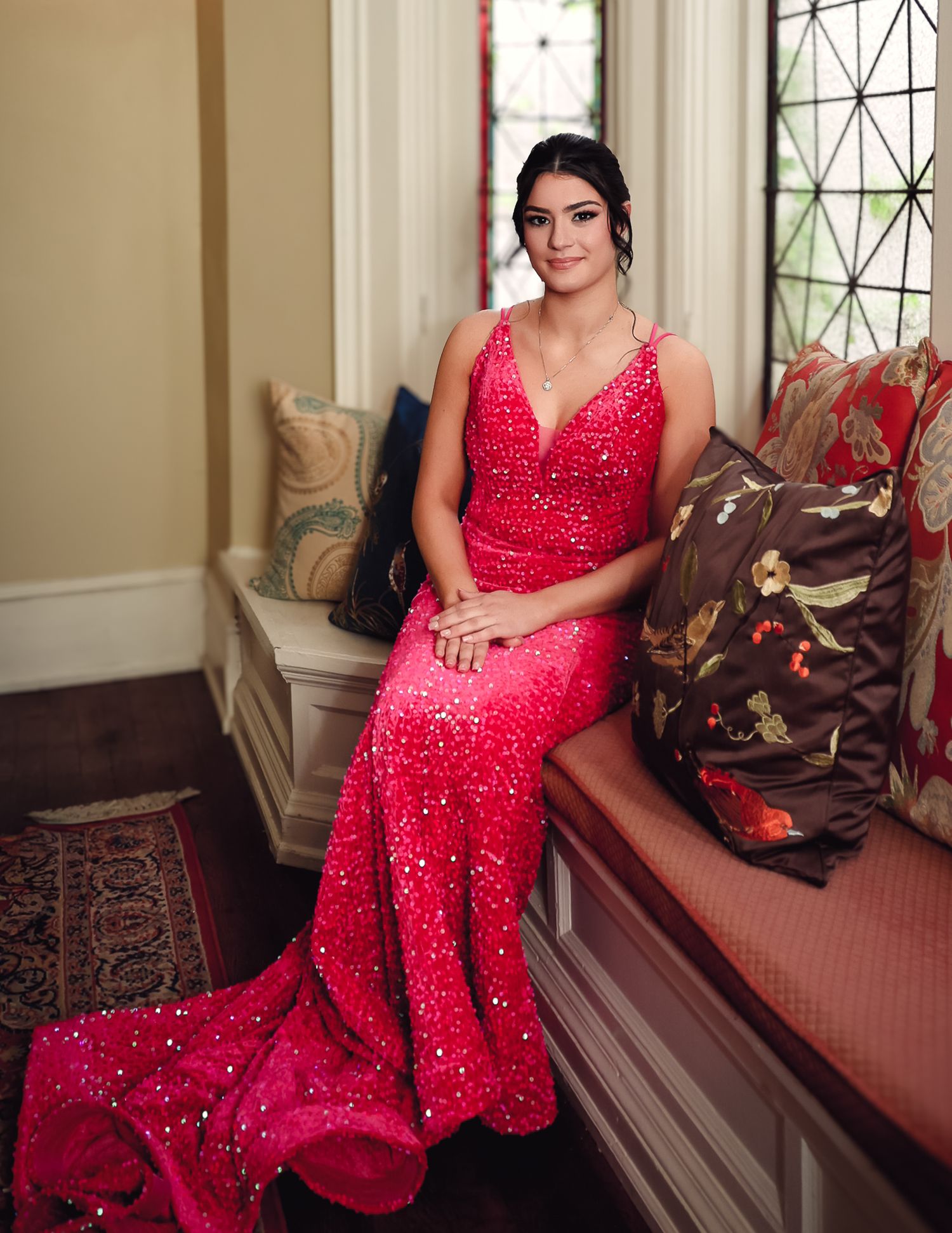 Young woman in a red sequined mermaid gown poses elegantly on a window seat with decorative pillows.