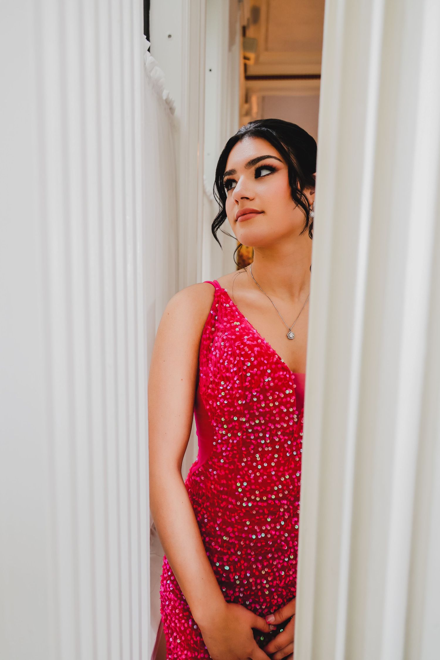 Young woman in a sparkling red sequin gown poses elegantly between white columns, wearing a delicate necklace.