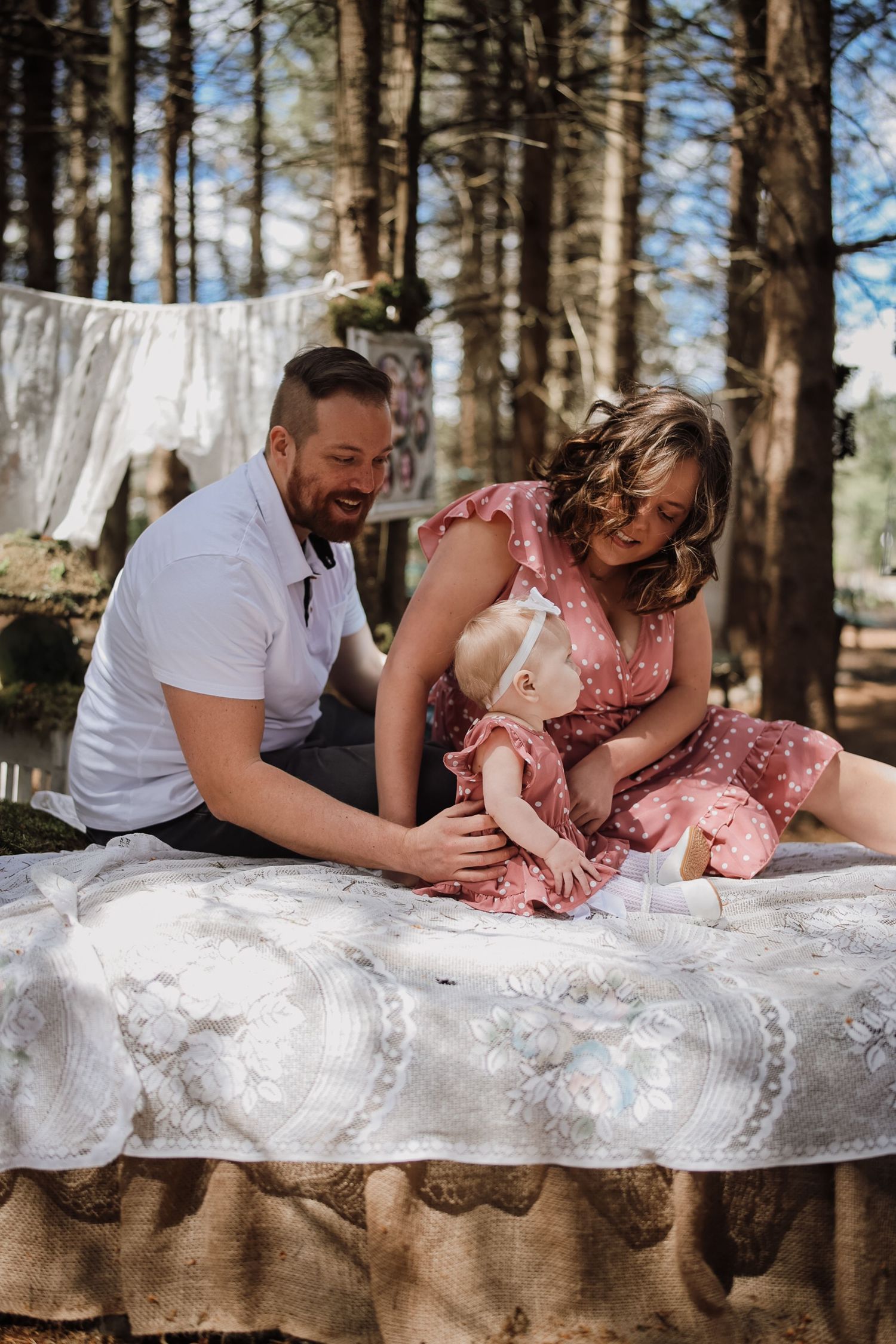 A couple in a forest setting smiles at their baby on a lace-covered surface, with laundry hanging in the background.