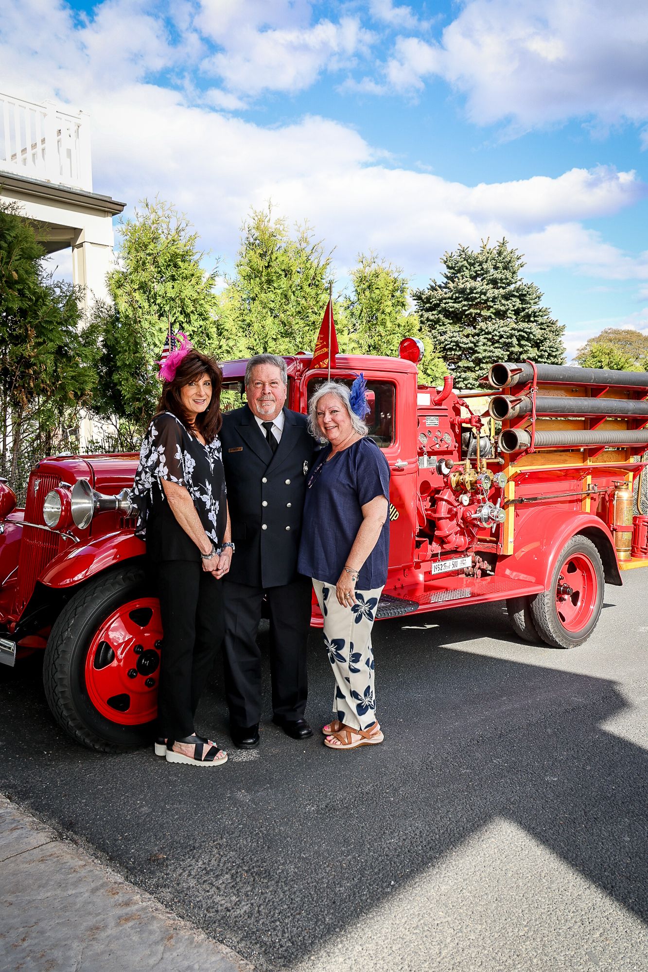 Three people pose proudly beside a vintage red fire truck outdoors on a sunny day.