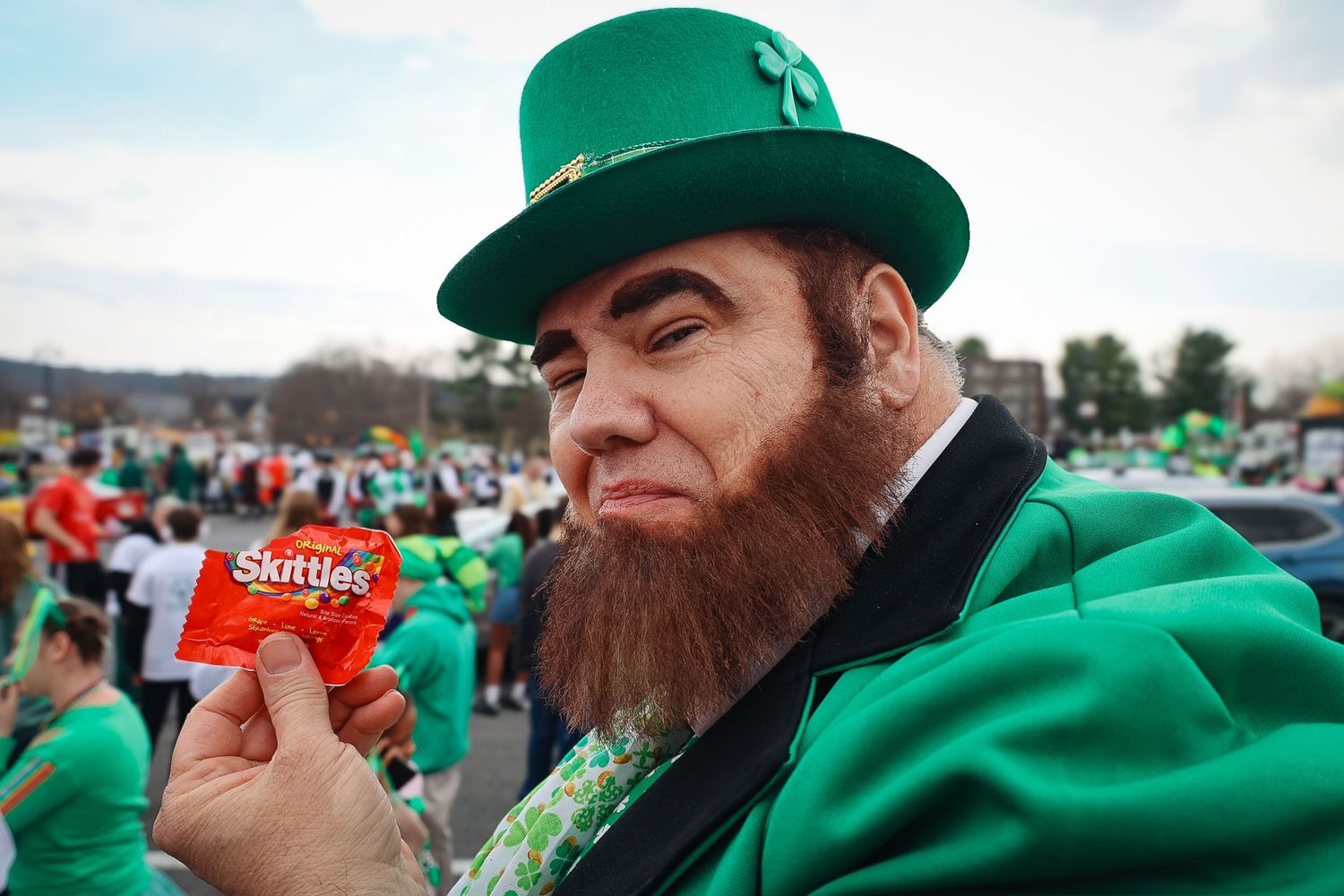 Man dressed as a leprechaun in green hat and suit holding Skittles candy at a St. Patrick's Day parade.
