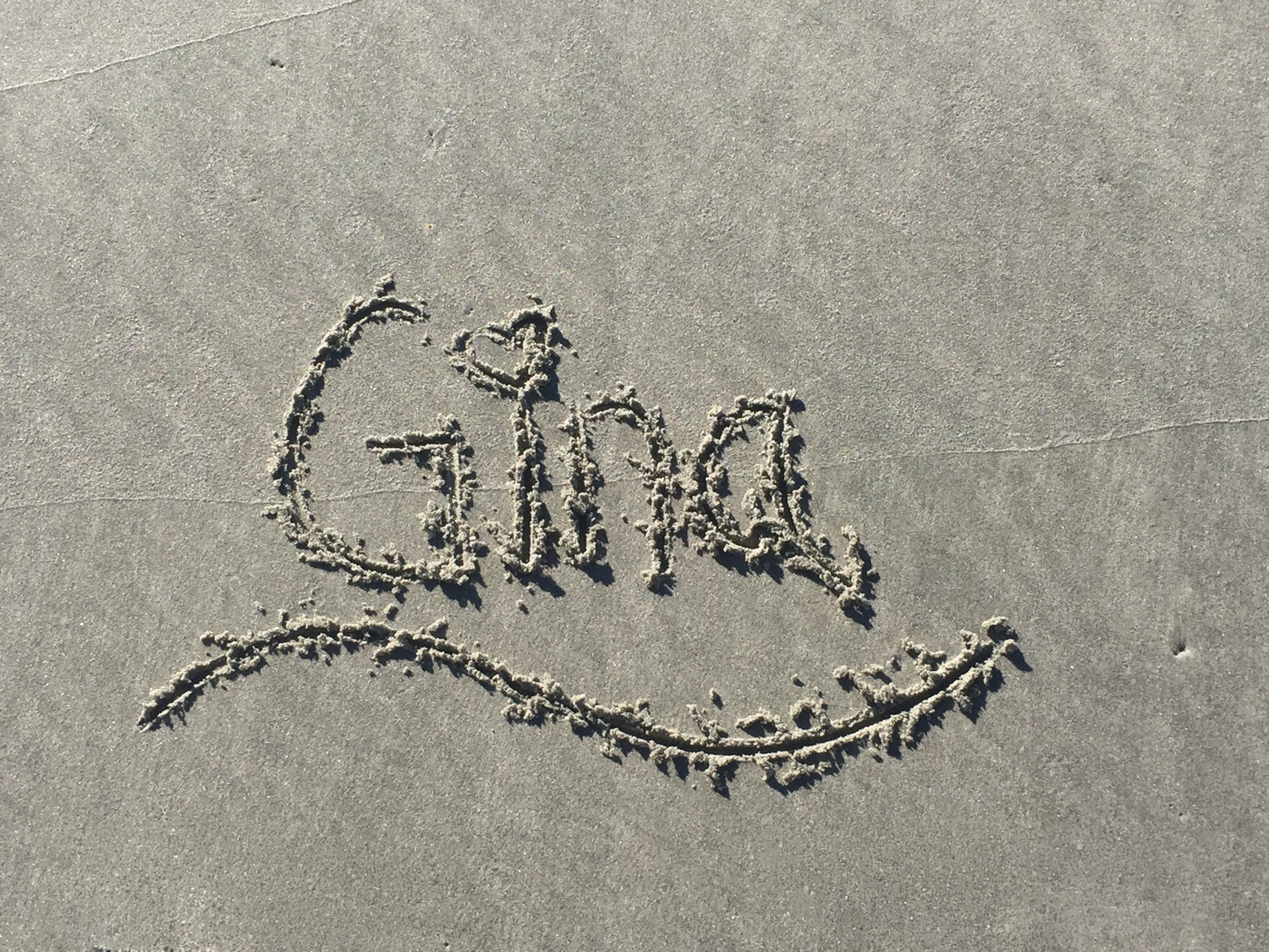 The name Gina written in sand on a beach with a heart and smiley face drawn around it.