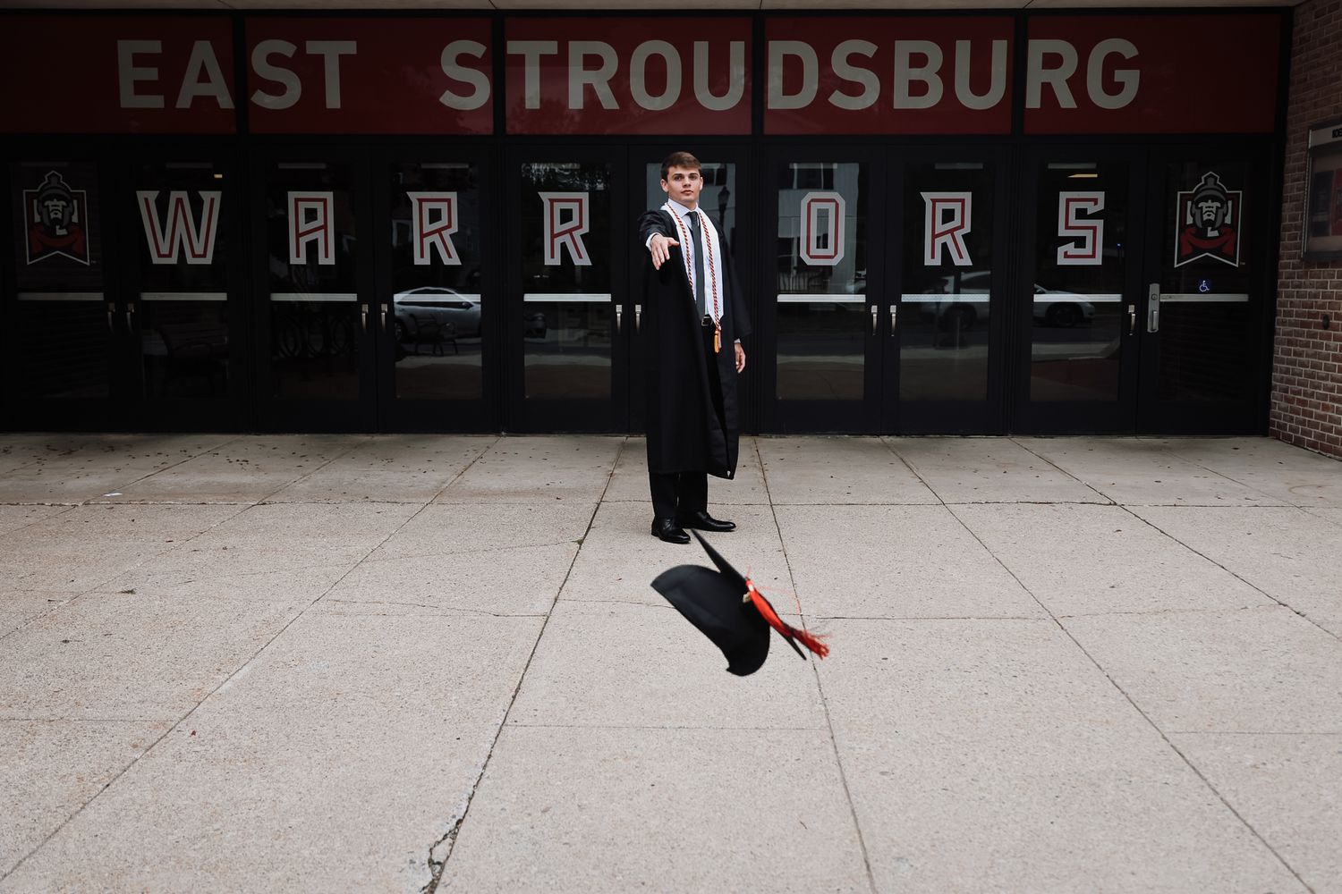 Graduate in cap and gown poses outside East Stroudsburg Warriors school building.