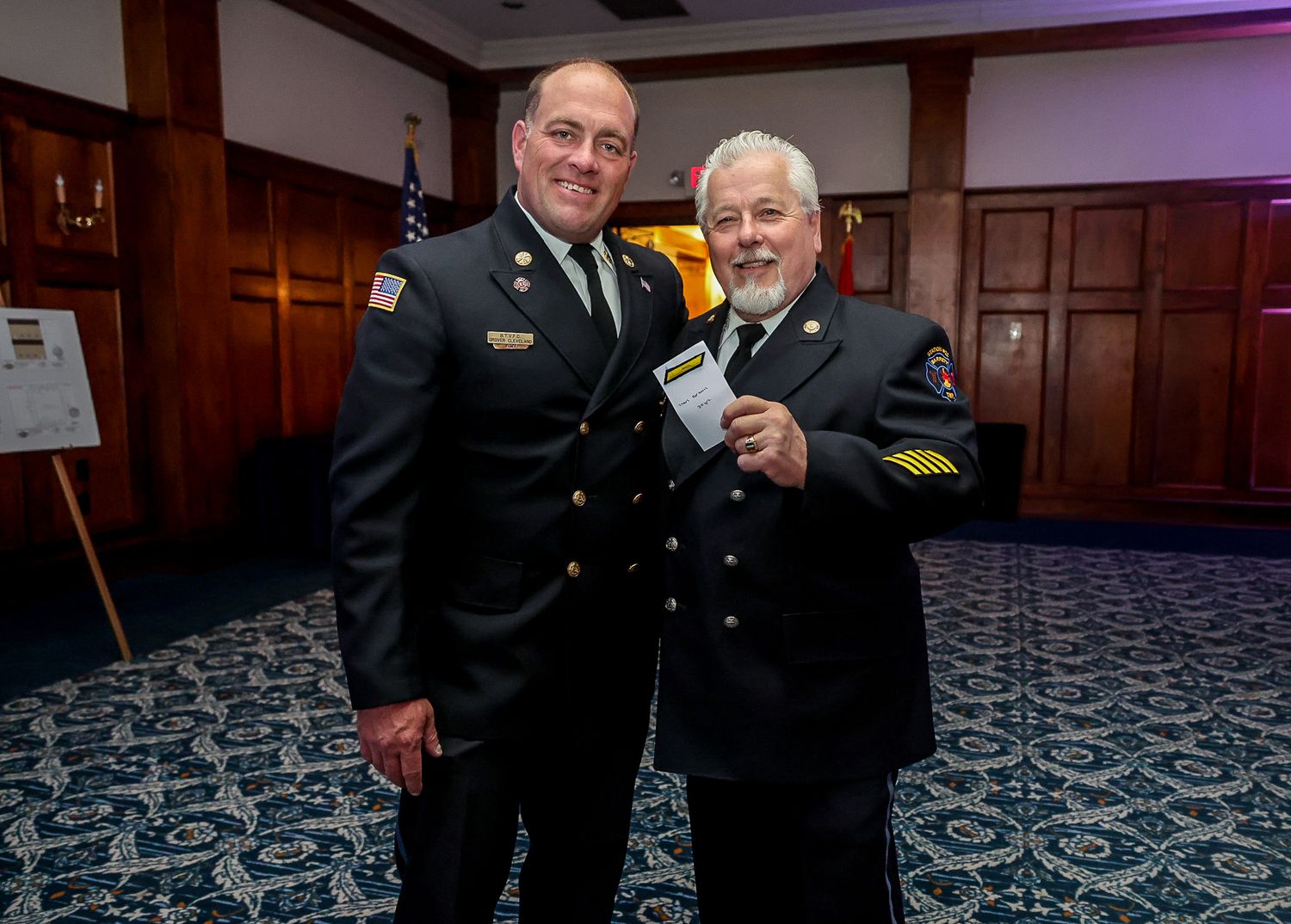Two firefighters in dress uniforms posing together at a formal ceremony, one holding an award certificate.