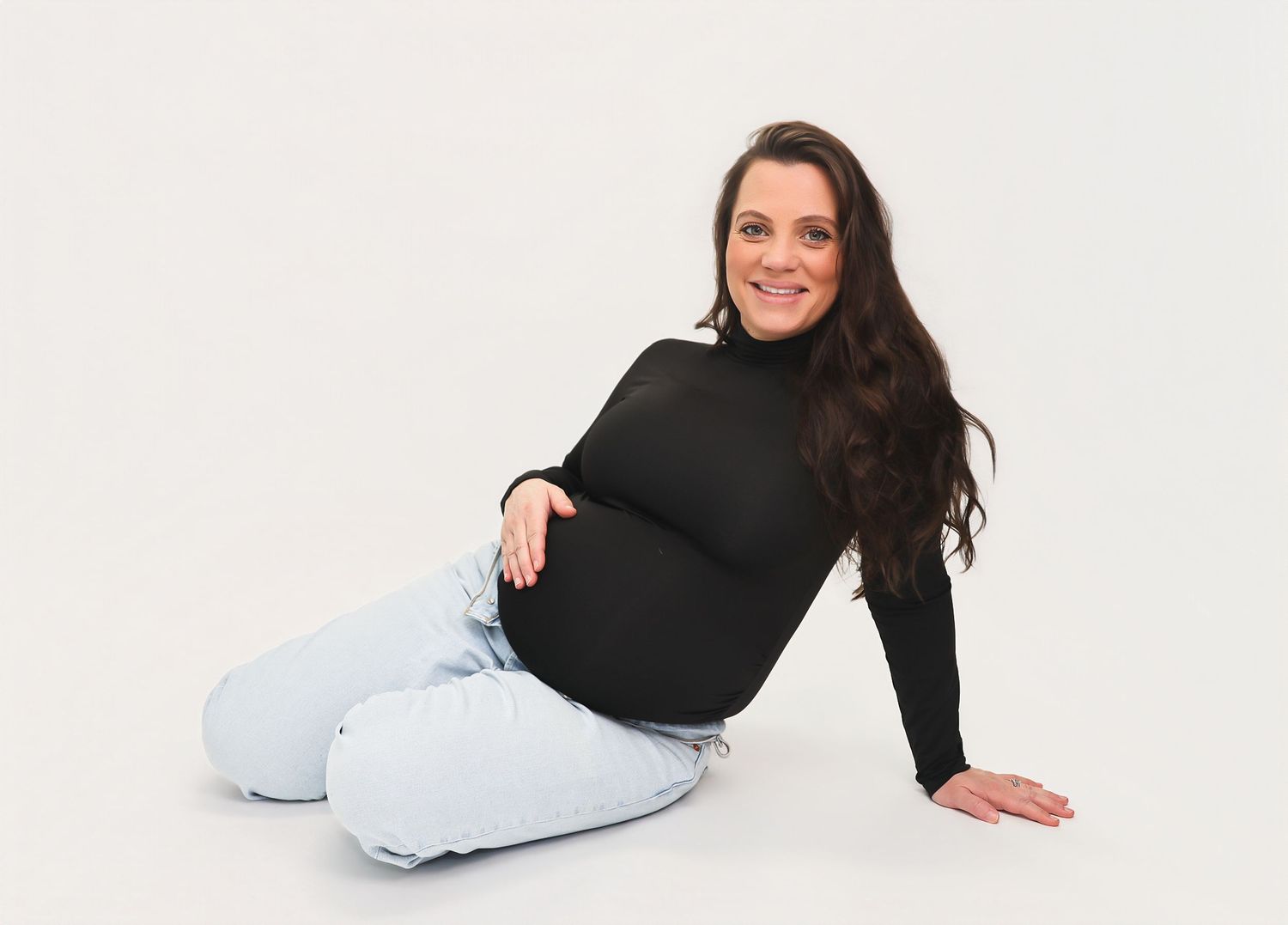 Smiling pregnant woman in black turtleneck and light jeans sitting on white background.