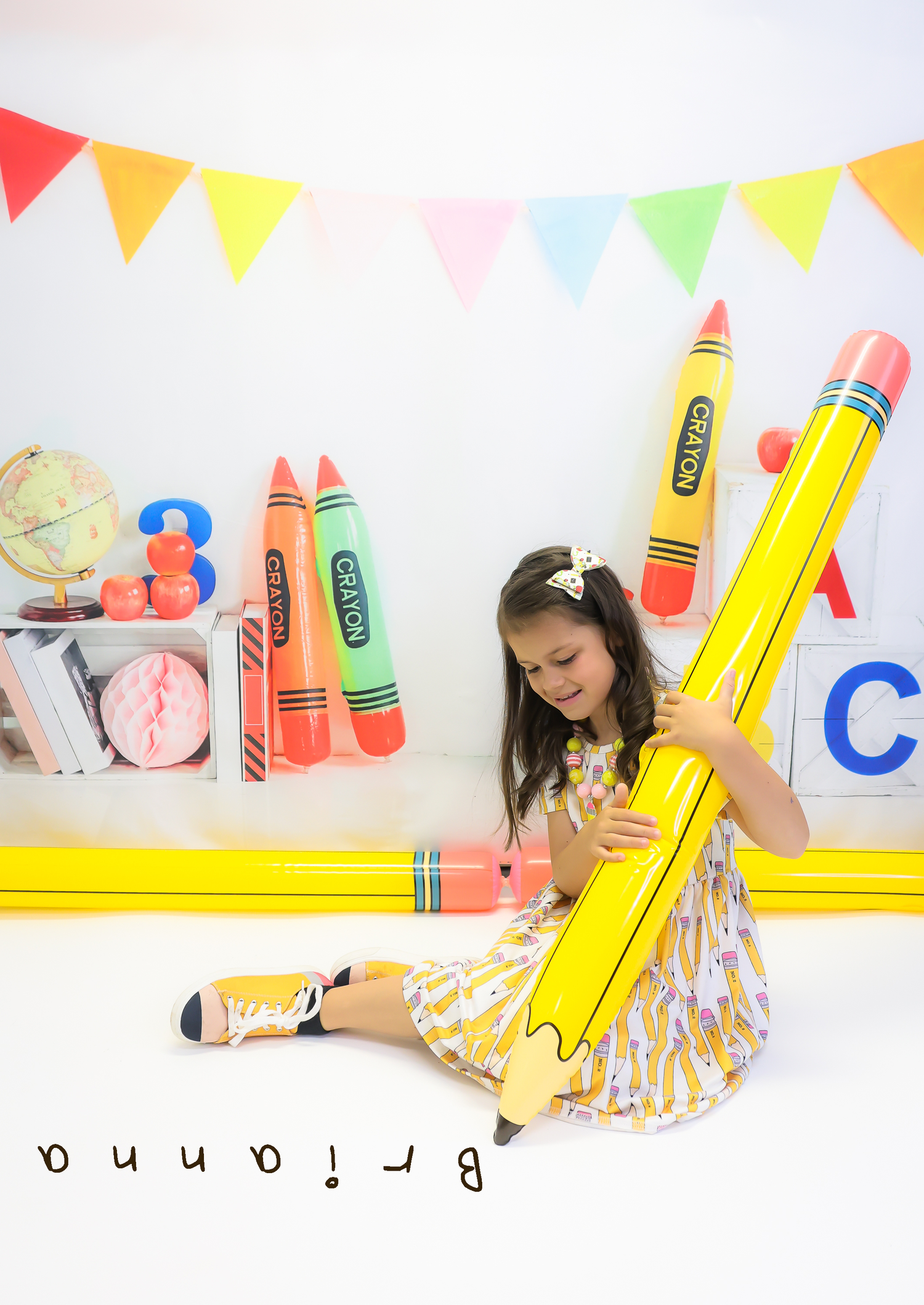 A young girl sits on the floor holding a giant inflatable yellow pencil surrounded by colorful crayon decorations and bunting.