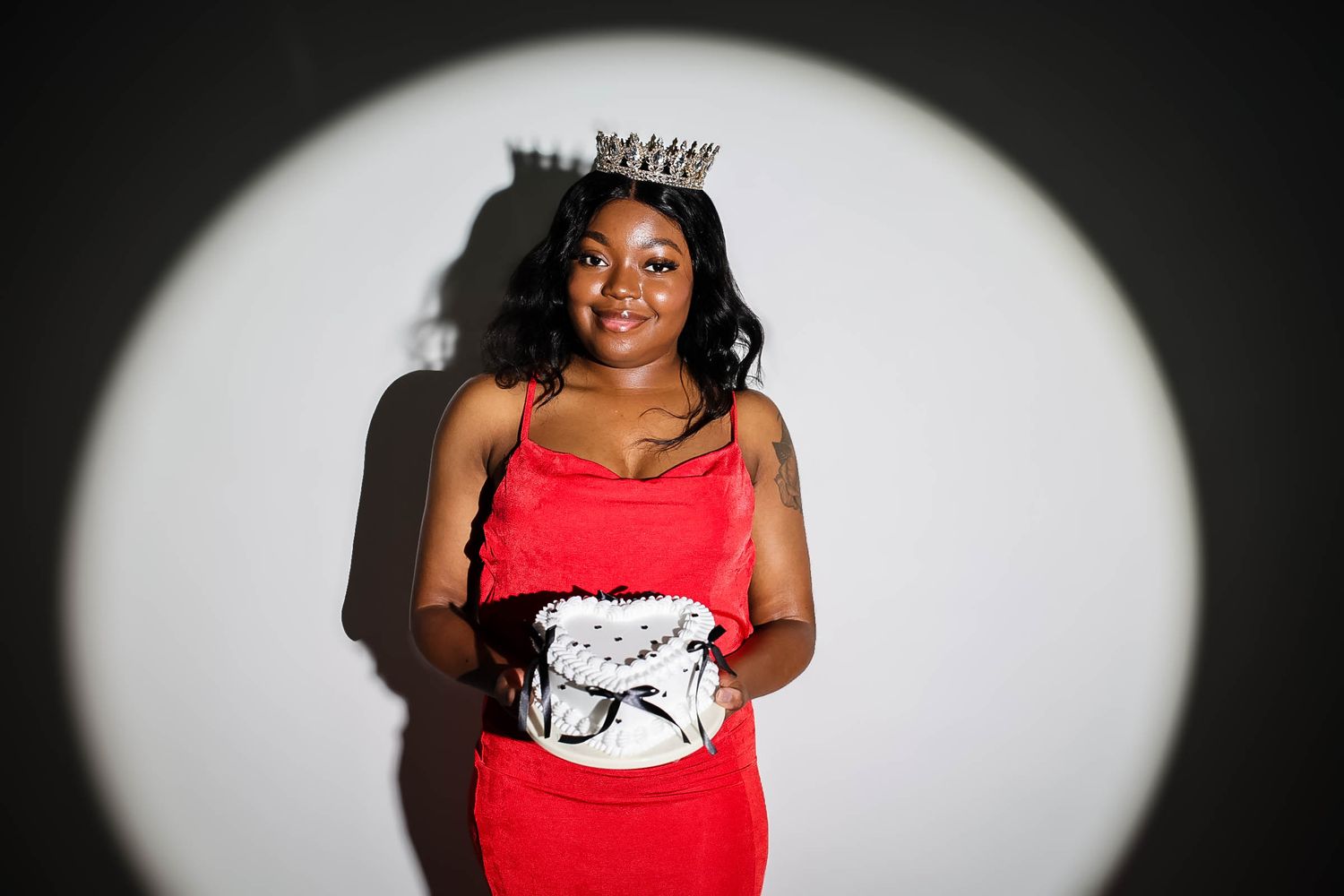 Young woman in red dress and silver crown poses confidently in spotlight against white background.