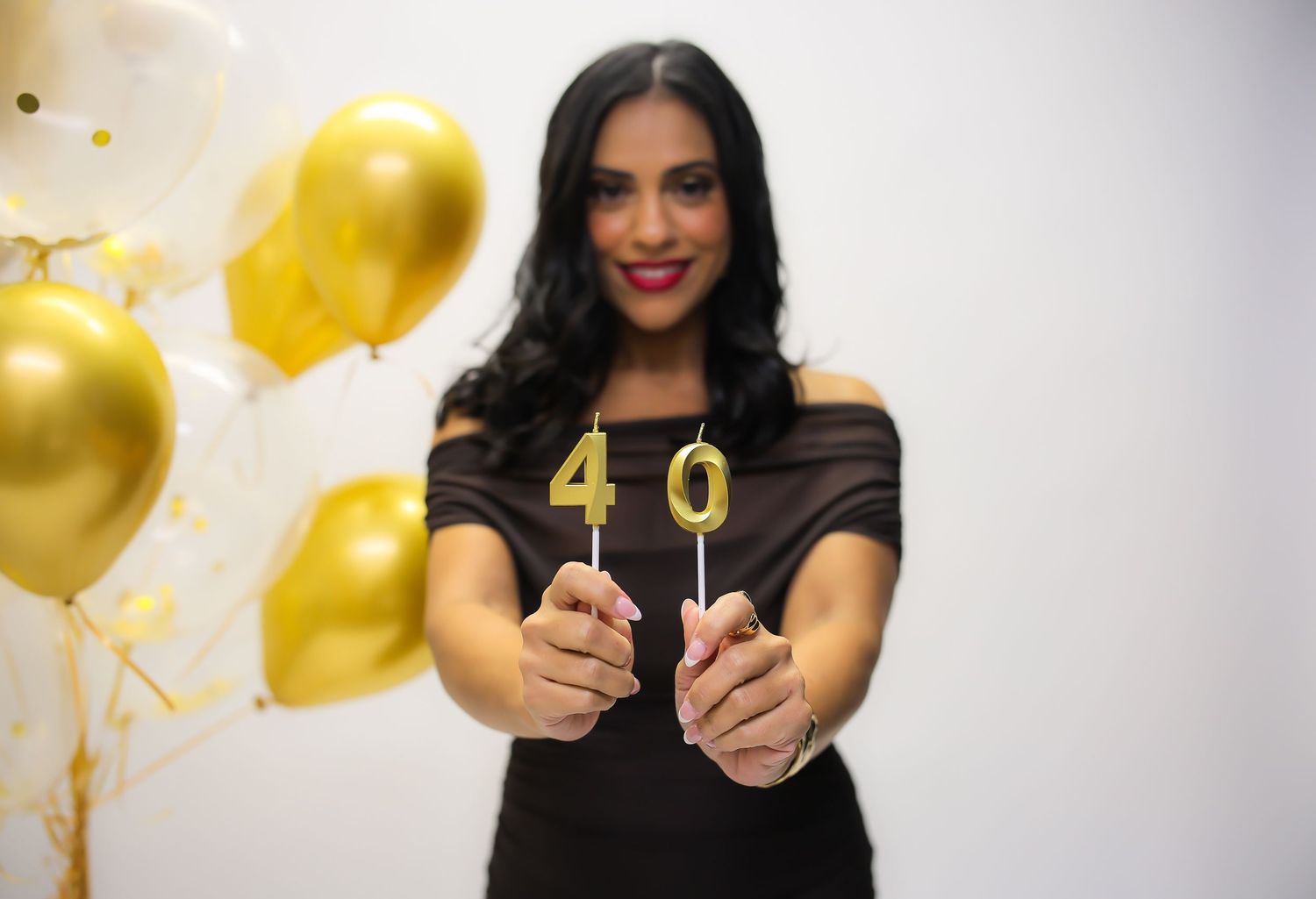 Woman in black dress holding gold number 40 candles, celebrating 40th birthday with gold and white balloons.