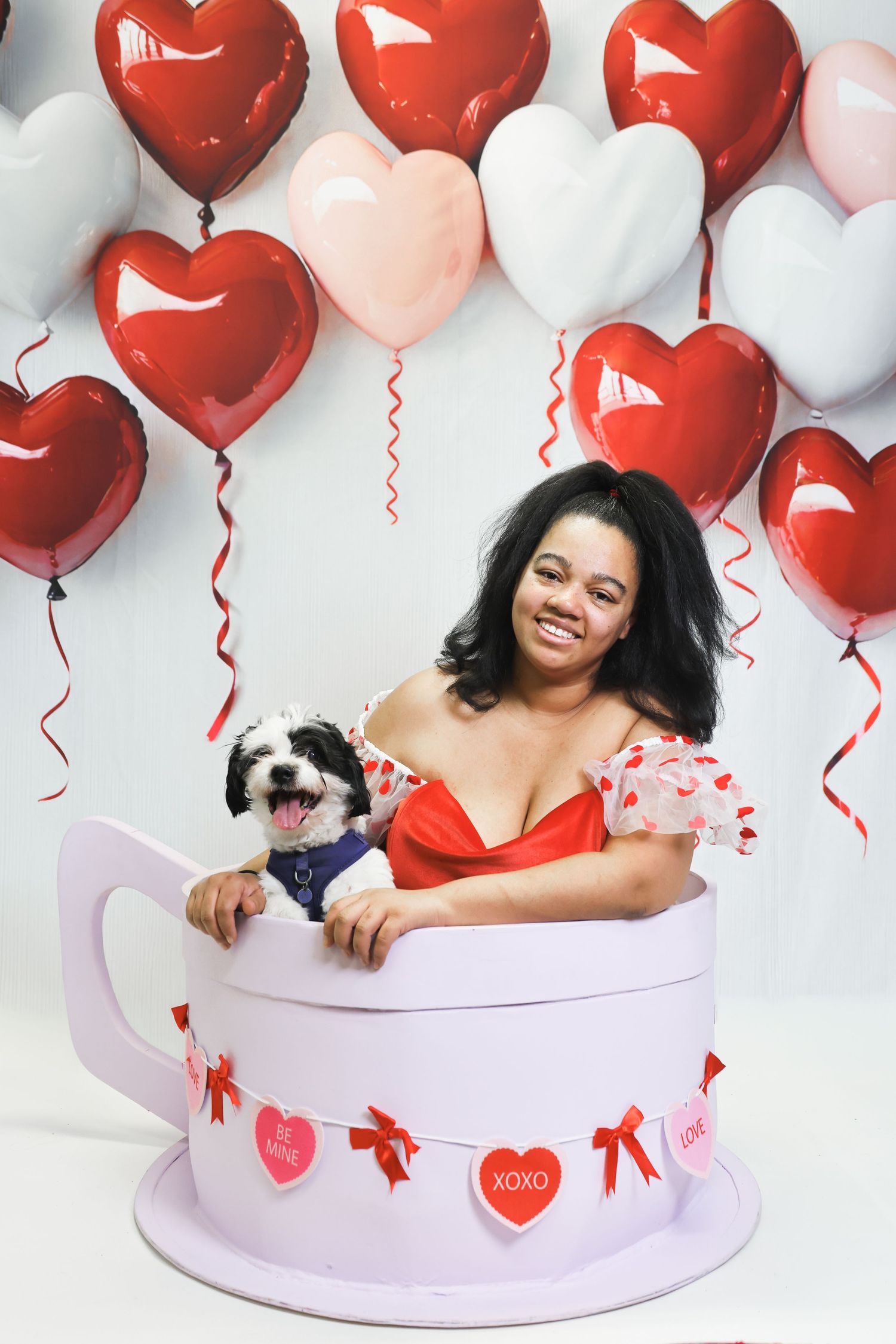 Smiling woman and fluffy dog pose in giant teacup with heart balloons for a festive Valentine's Day photo shoot.