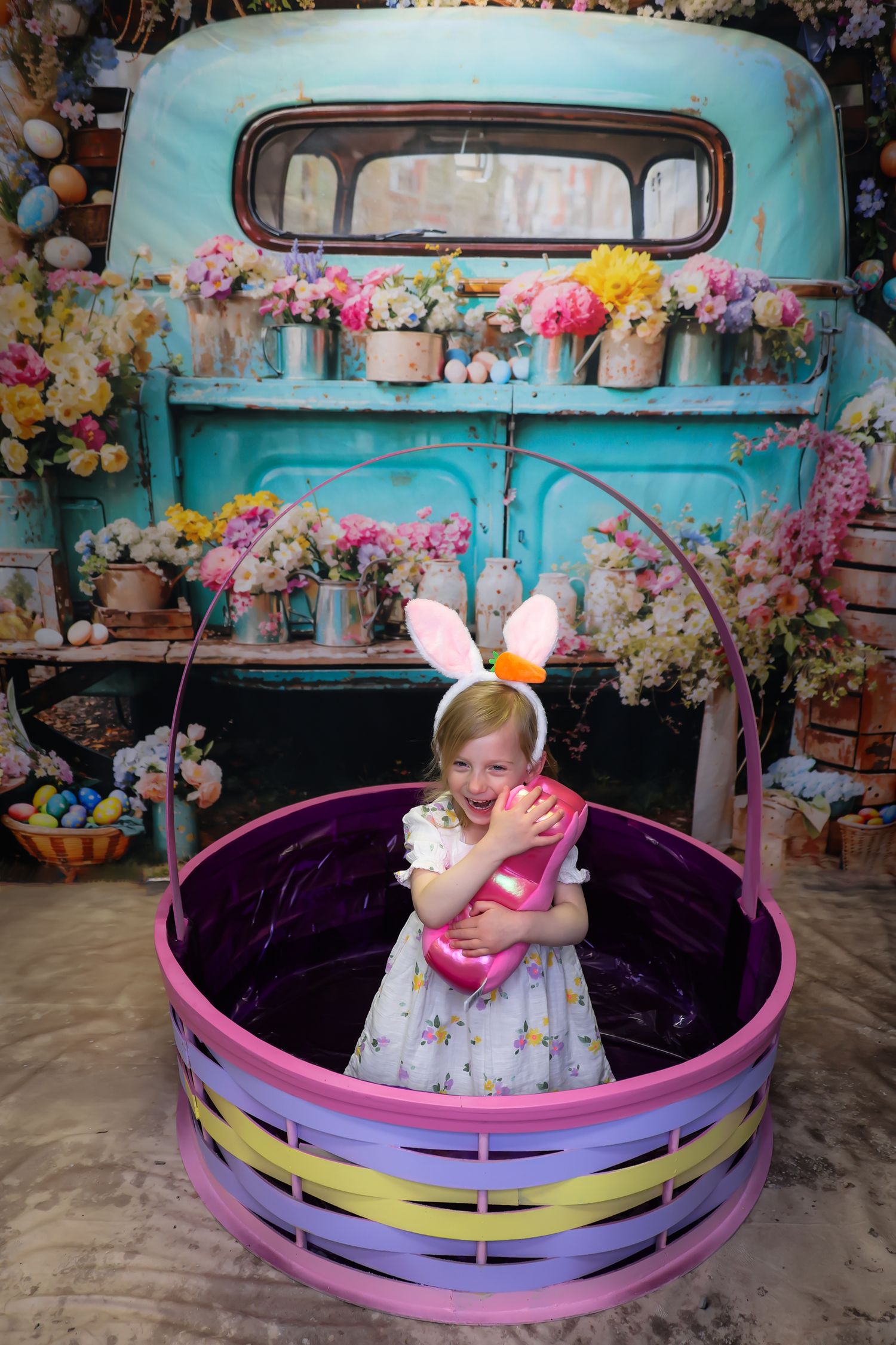Young girl in bunny ears sits in large Easter basket prop before vintage teal truck backdrop with colorful spring flowers.