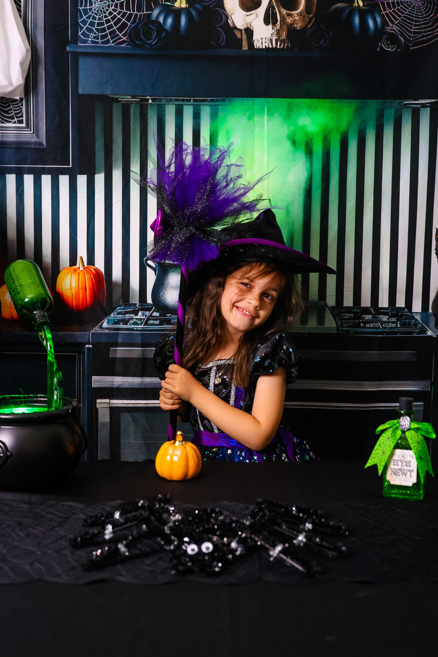 Young girl in witch costume with purple hat smiles at Halloween decorated table with pumpkins and cauldron.
