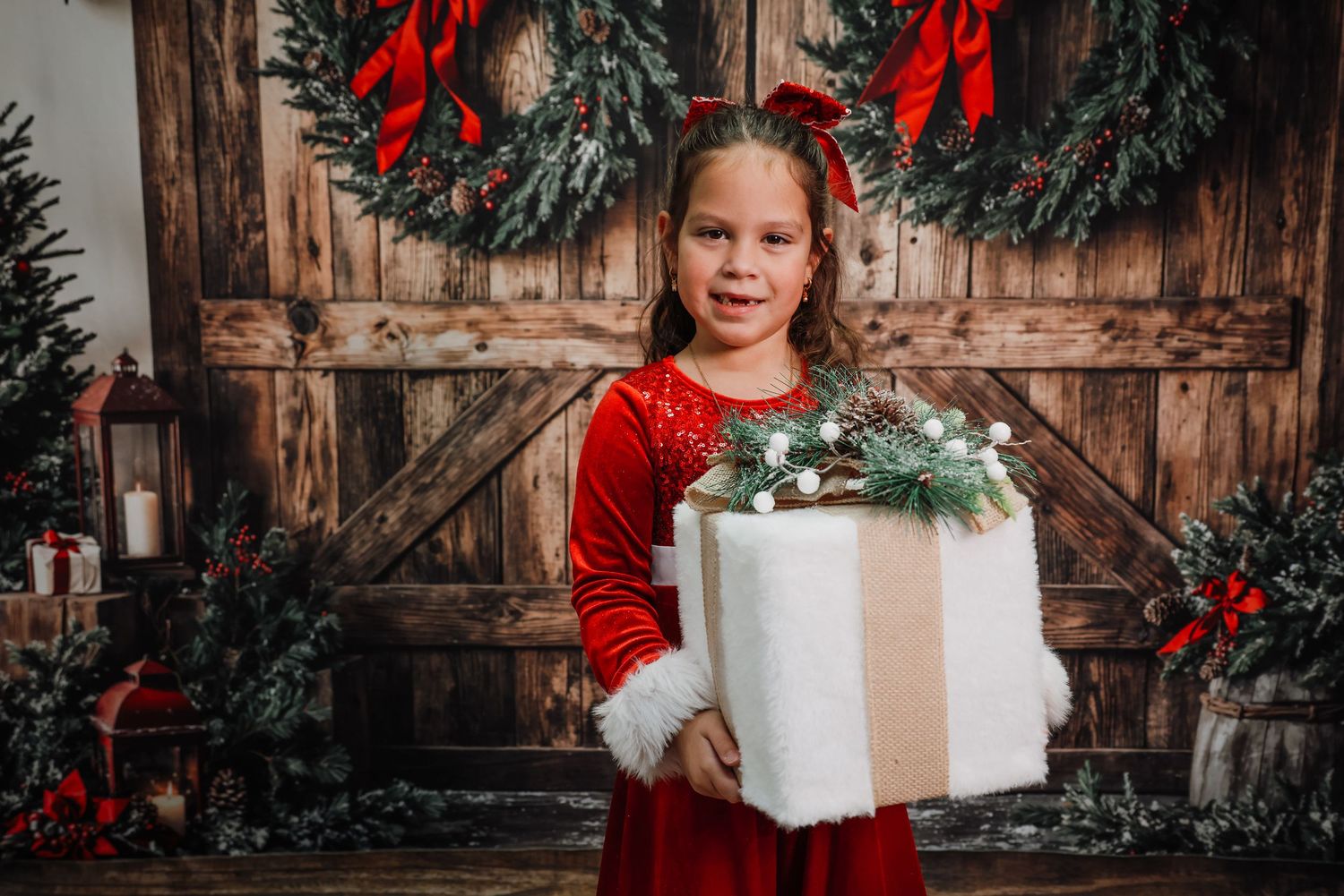 Young girl in red Christmas dress holds large gift box in front of festive wooden door with holiday wreaths.