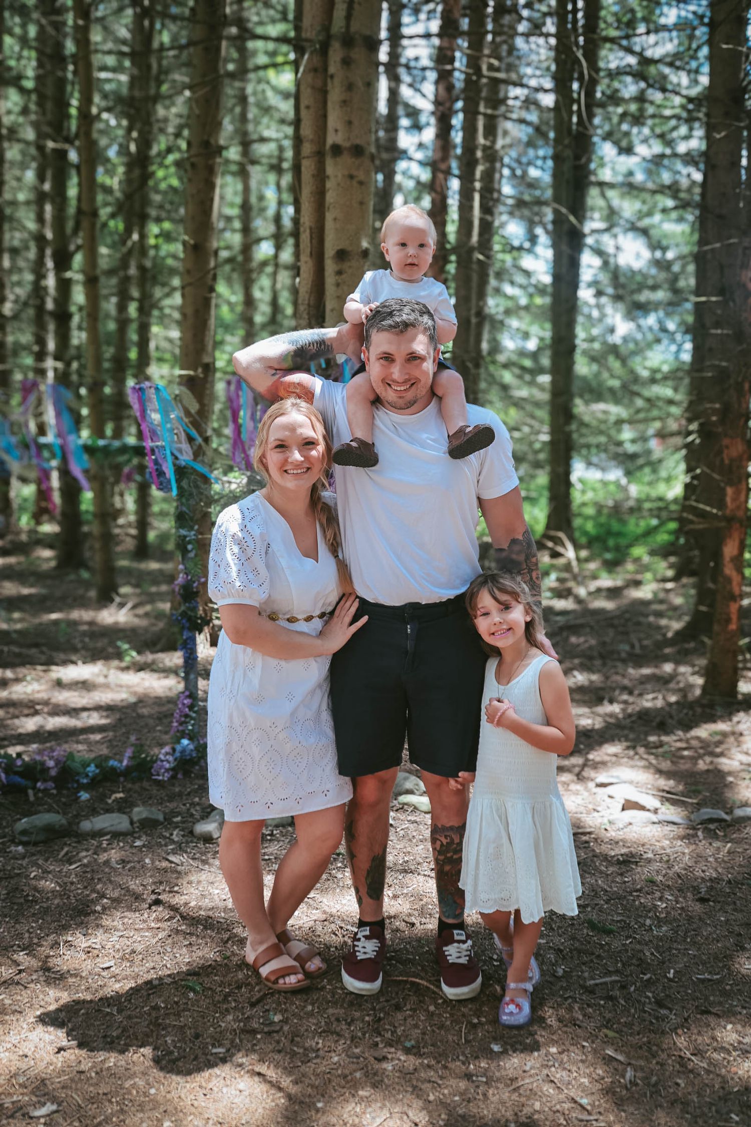 Family of four posing in a forest, parents in white outfits with two young children, baby on dad's shoulders.