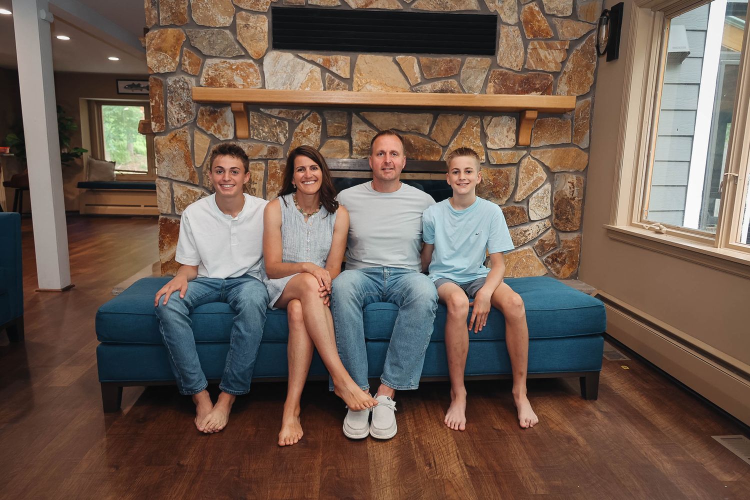 Family of four smiling together on a blue couch in a cozy stone fireplace living room.