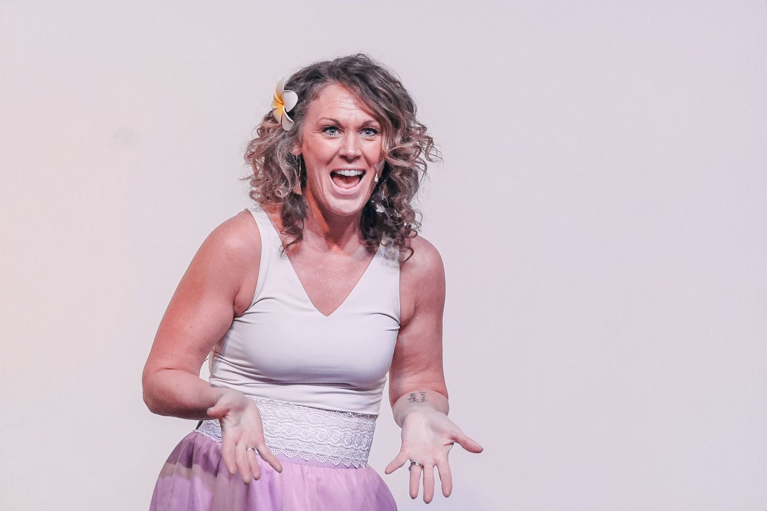 Woman in white top and purple skirt laughing expressively with hands outstretched against light background.