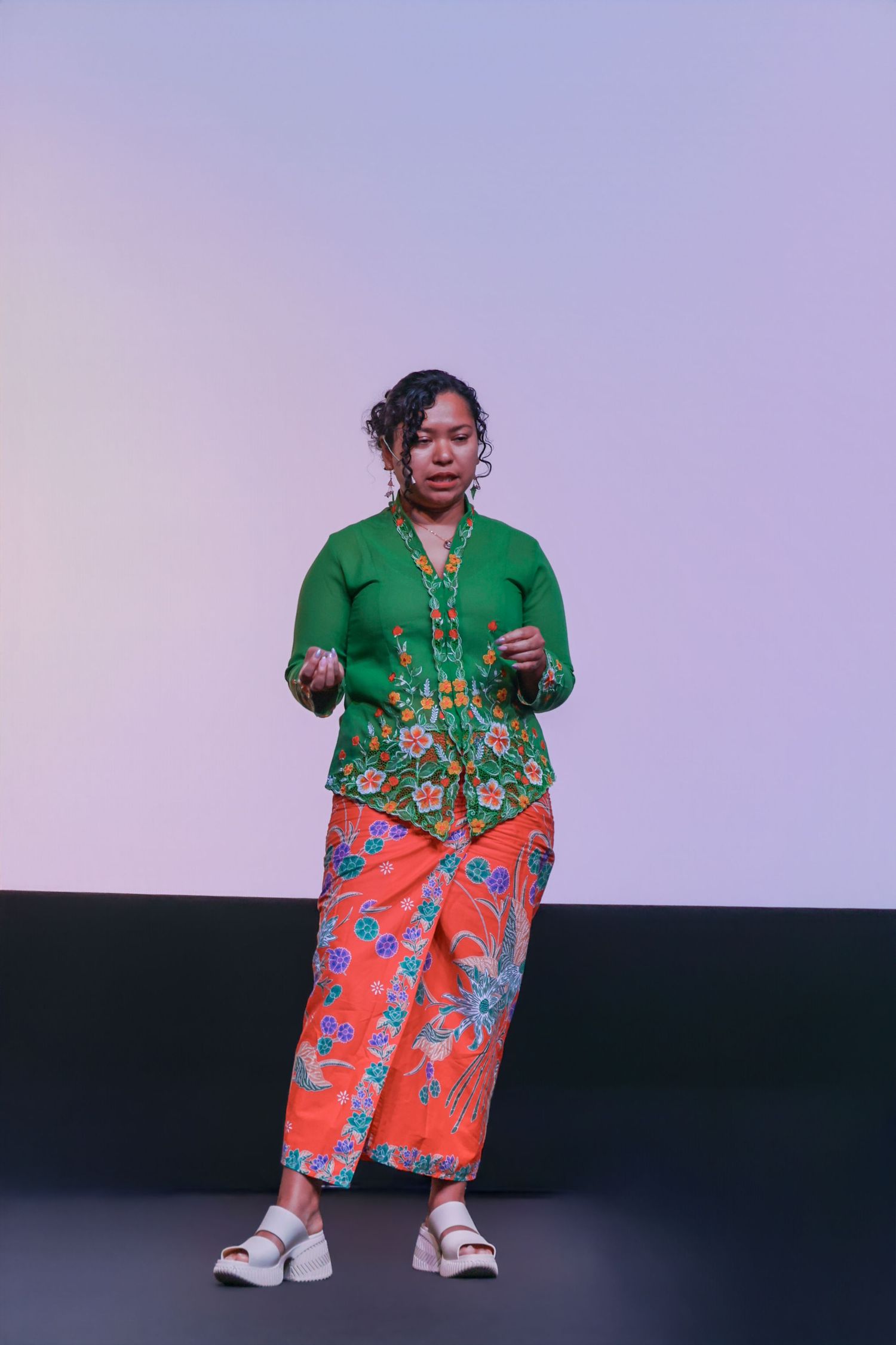 Woman in green floral top and orange batik skirt posing on stage in traditional Southeast Asian attire.