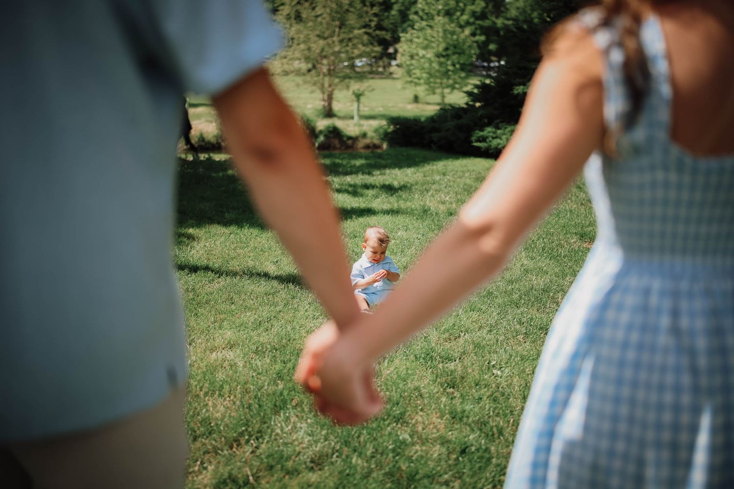 Couple holding hands in a sunny park while a young child stands in the background on green grass.
