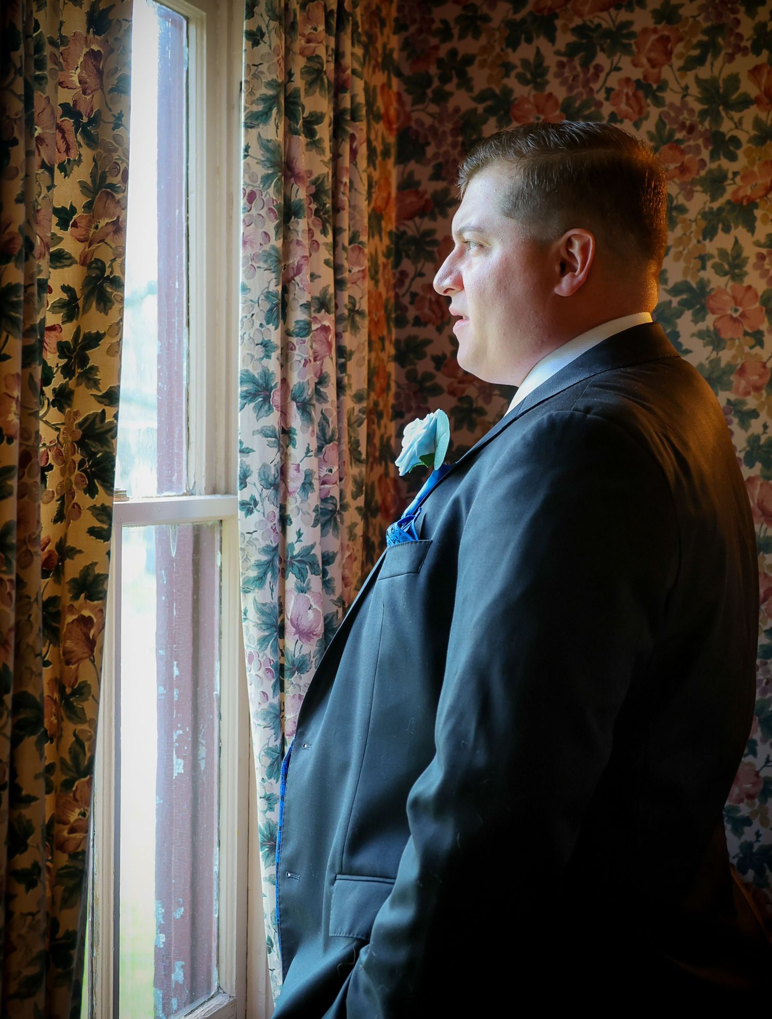 A groom in a black suit with a blue boutonniere gazes thoughtfully out a window beside floral curtains.