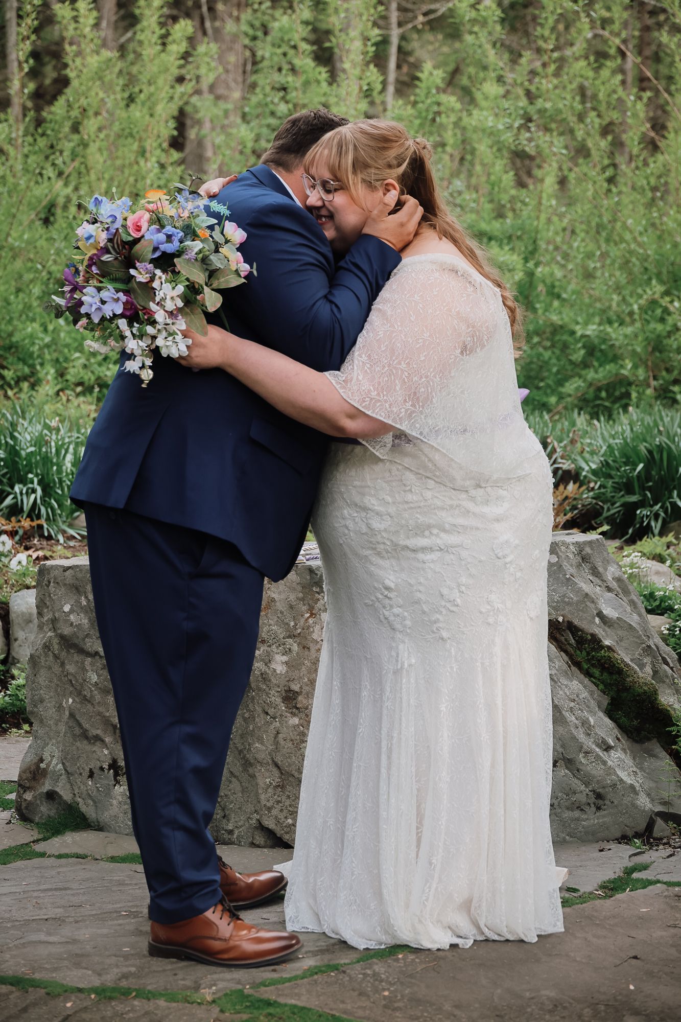 Bride and groom share first kiss in garden wedding, bride in lace gown holding colorful bouquet, groom in navy suit.