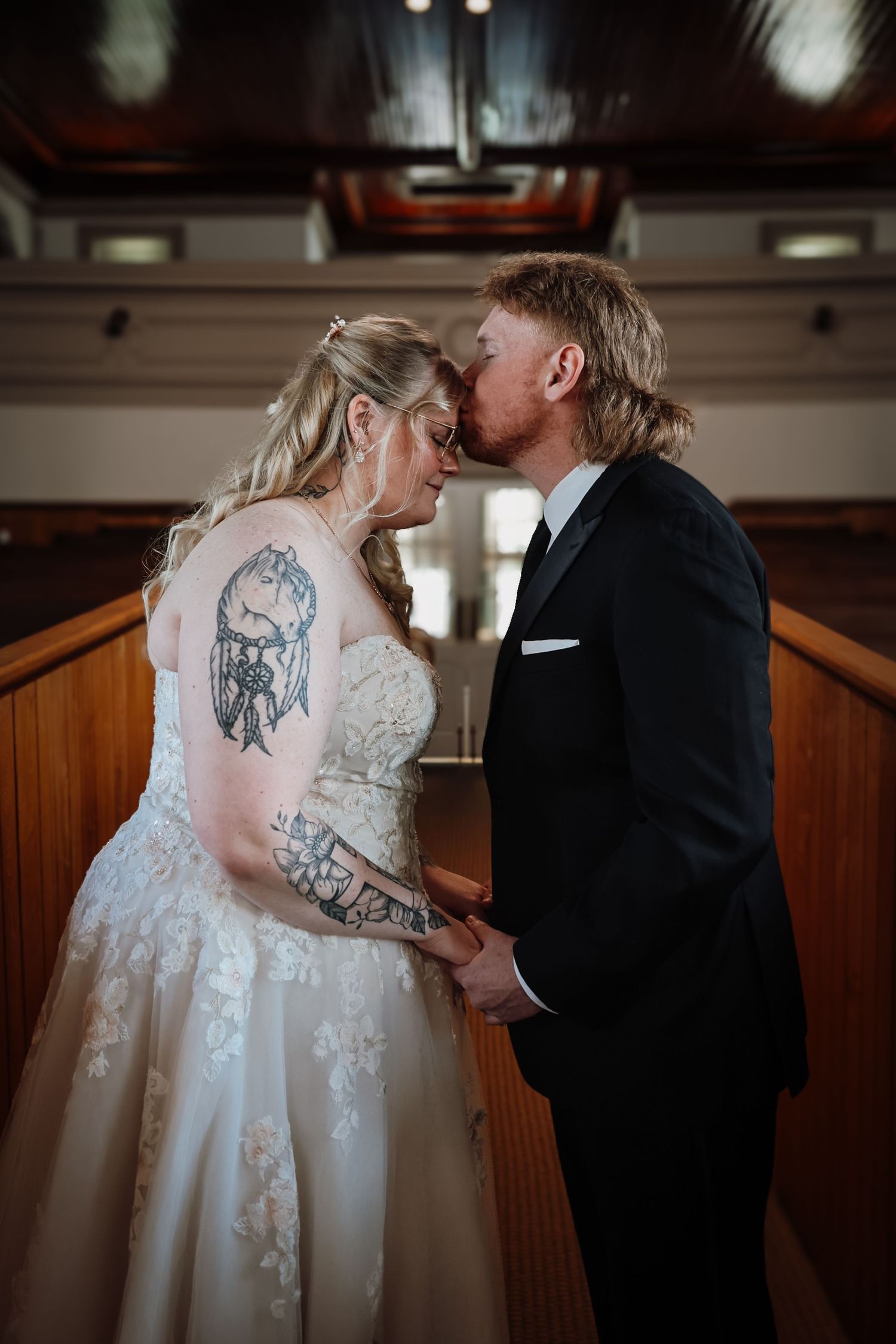 Bride and groom share a kiss at the altar, bride wearing lace gown with tattoos, groom in black suit.
