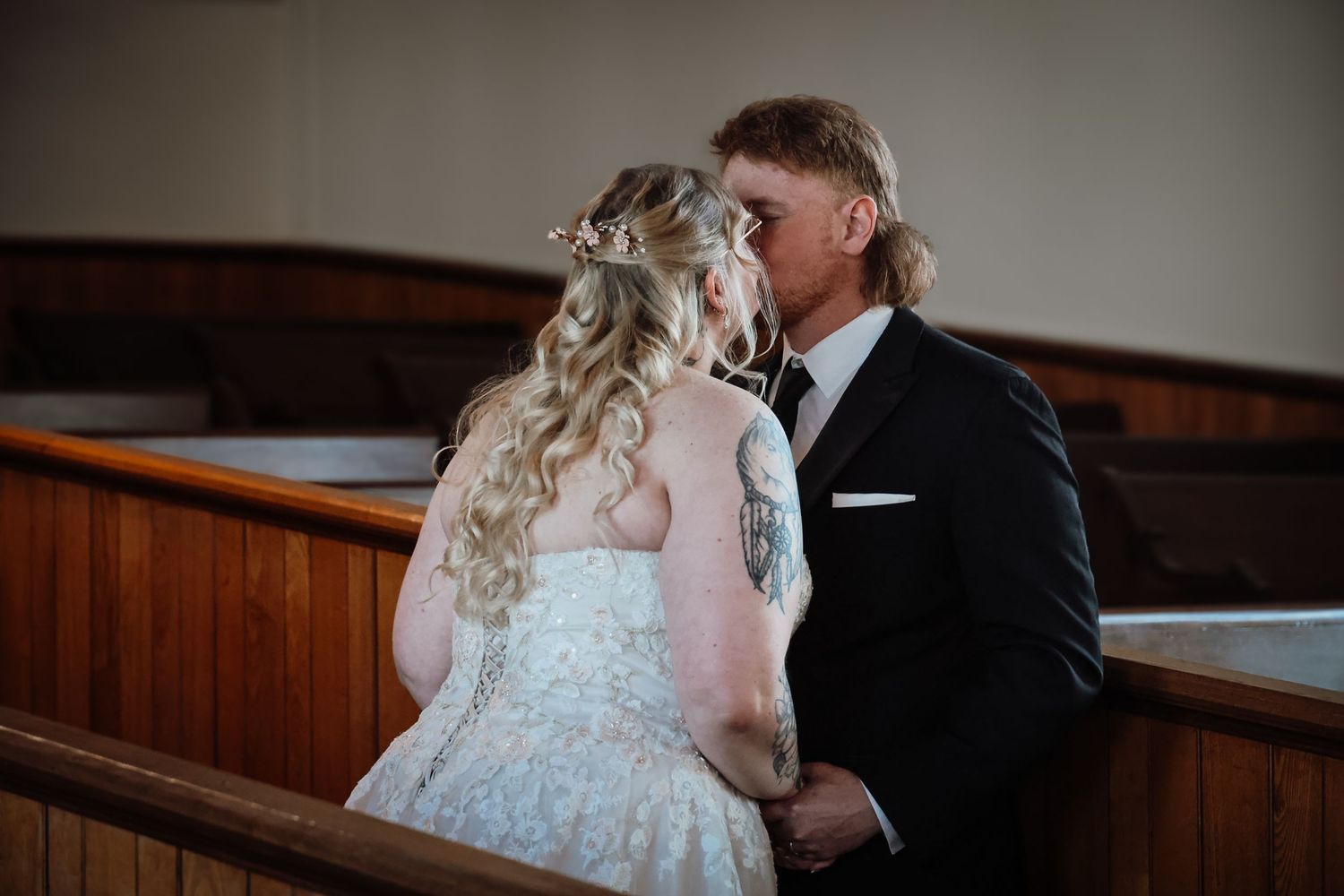 A bride and groom share a tender kiss inside a church, with the bride in a lace gown and groom in a black suit.