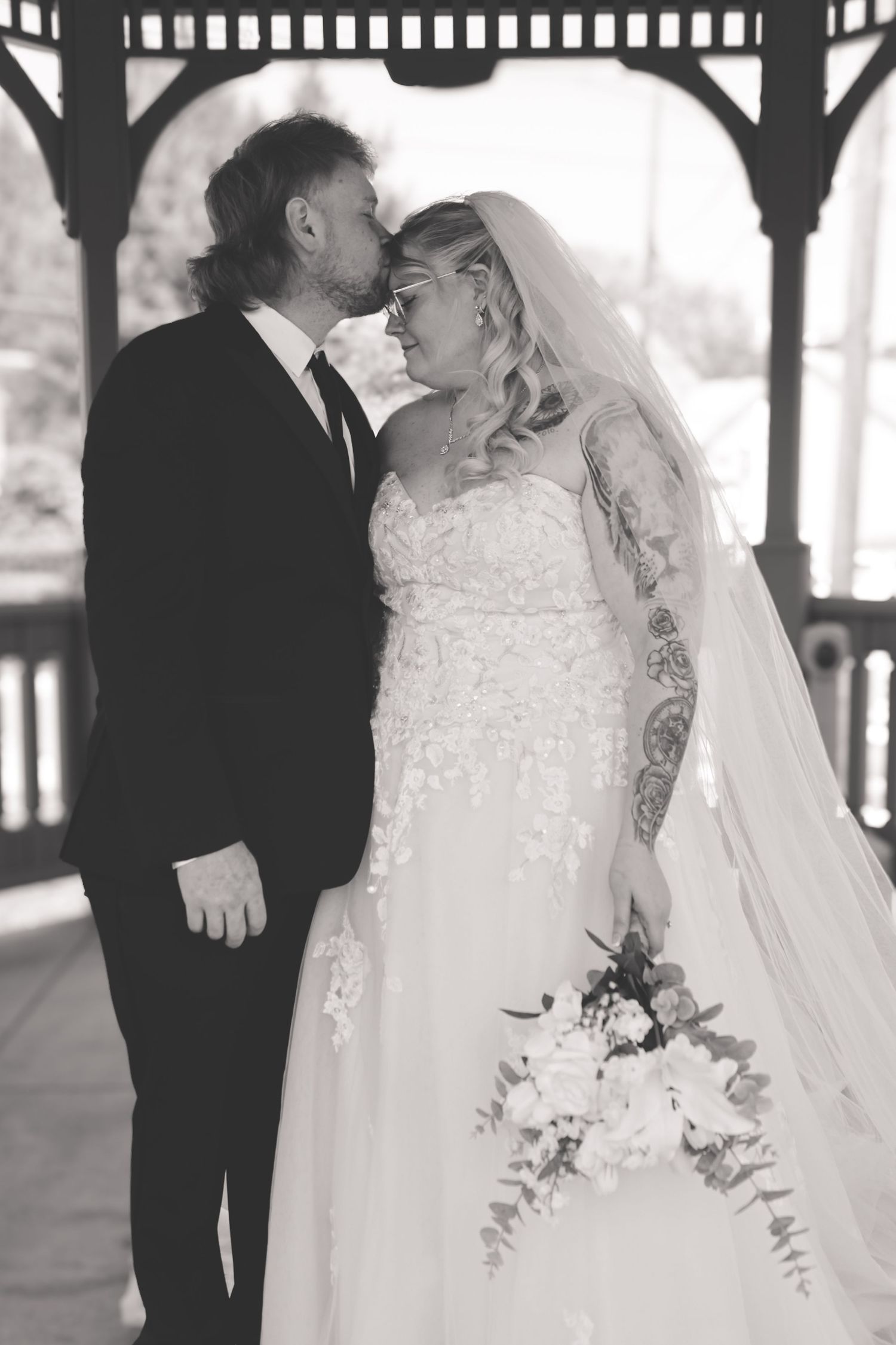 Black and white wedding photo of bride and groom sharing a kiss under a gazebo, bride holding a floral bouquet.