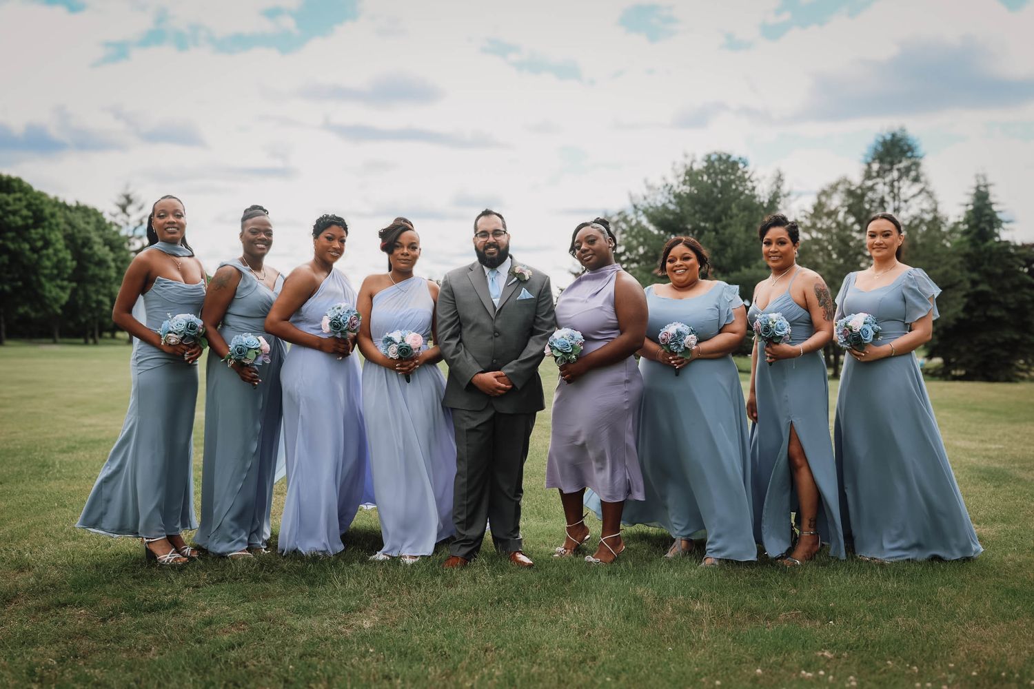 Groom in gray suit stands with eight bridesmaids in light blue dresses holding bouquets on a green lawn.