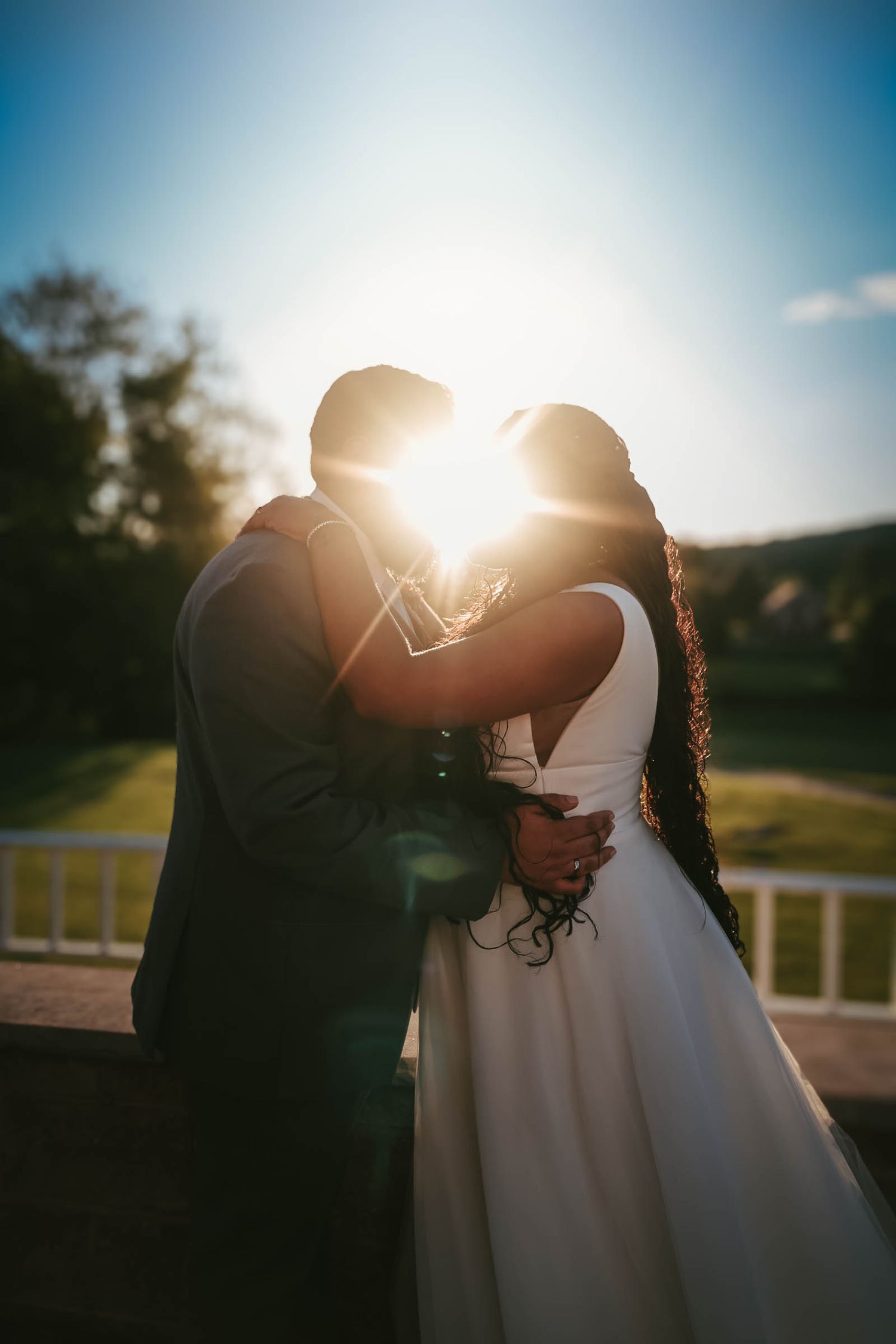 A couple shares a romantic kiss at sunset, with golden light glowing behind them on their wedding day.