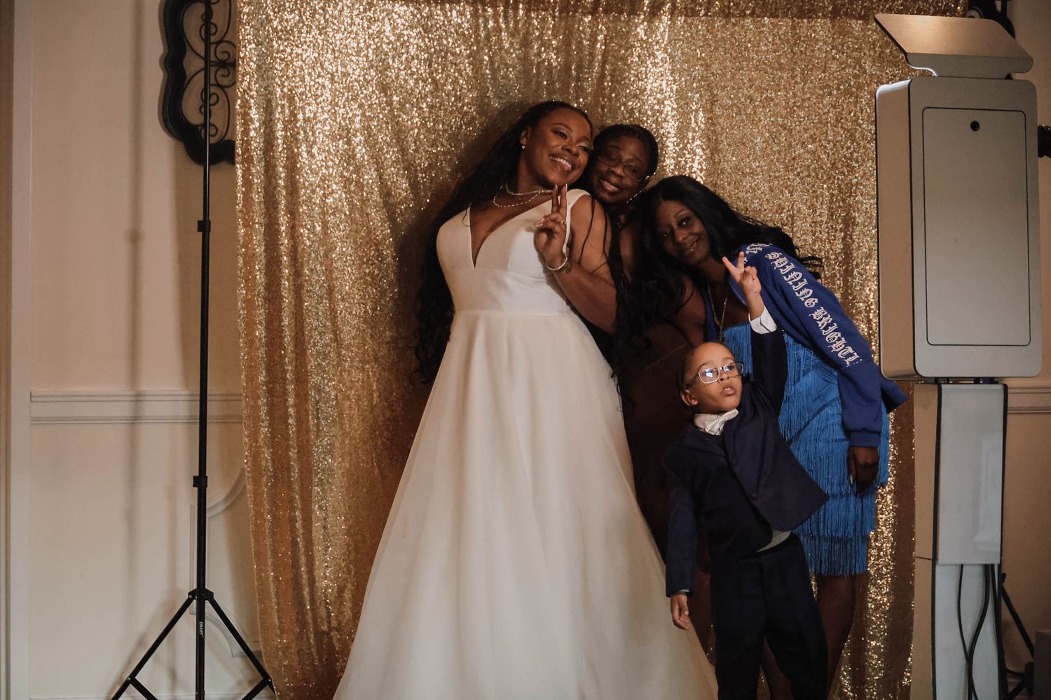 Bride in white gown poses joyfully with three guests in front of gold sequin photo booth backdrop at wedding reception.