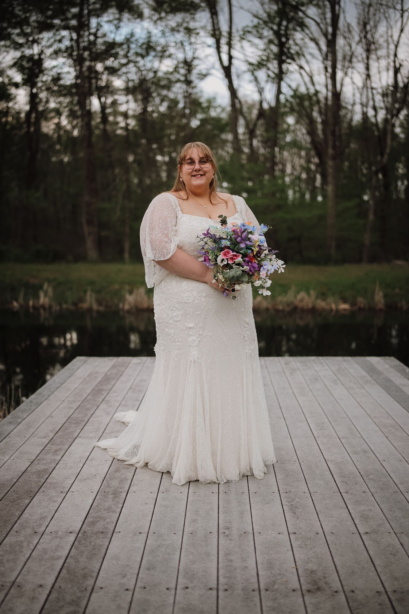A bride in a lace wedding gown holds a colorful bouquet on a wooden dock surrounded by trees and a serene pond.