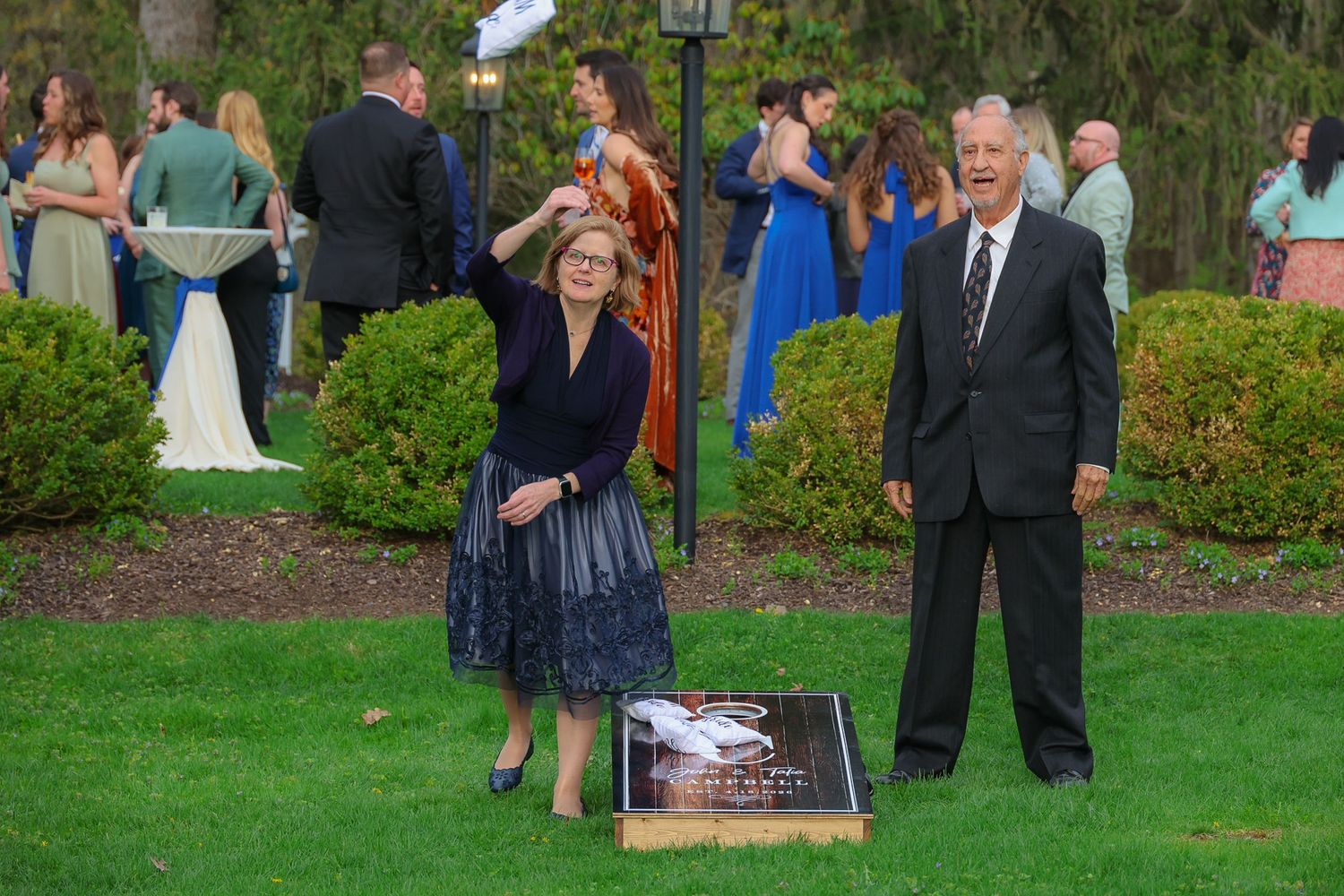 Couple playing cornhole lawn game at outdoor wedding reception on green grass with guests in background.