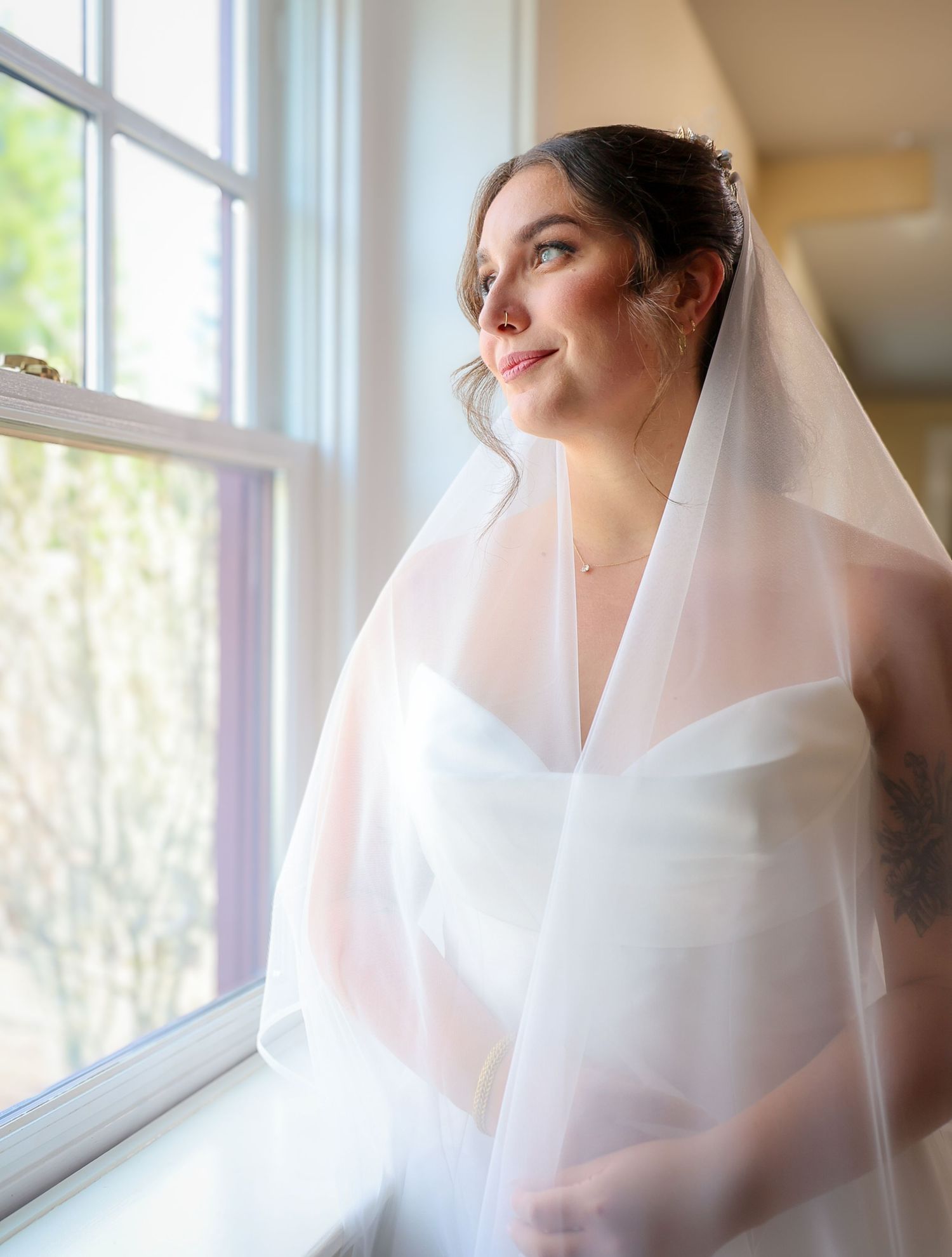 Bride in white strapless gown and veil gazes out sunlit window, showcasing elegant bridal look.