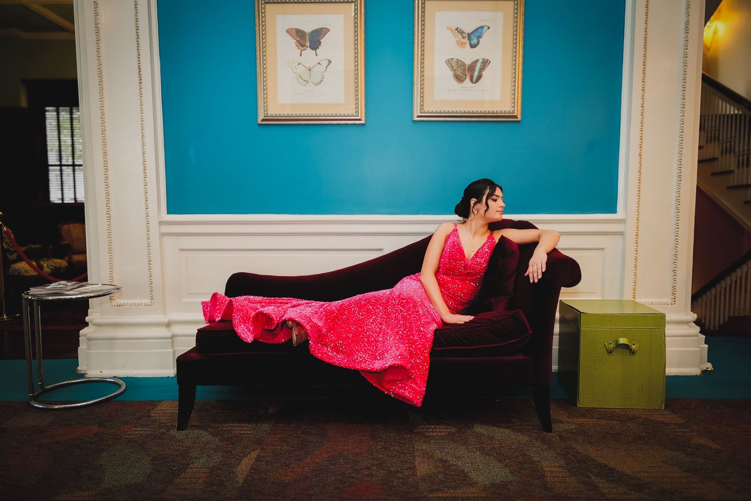Woman in red gown reclining on velvet chaise lounge in elegant teal room with butterfly artwork.