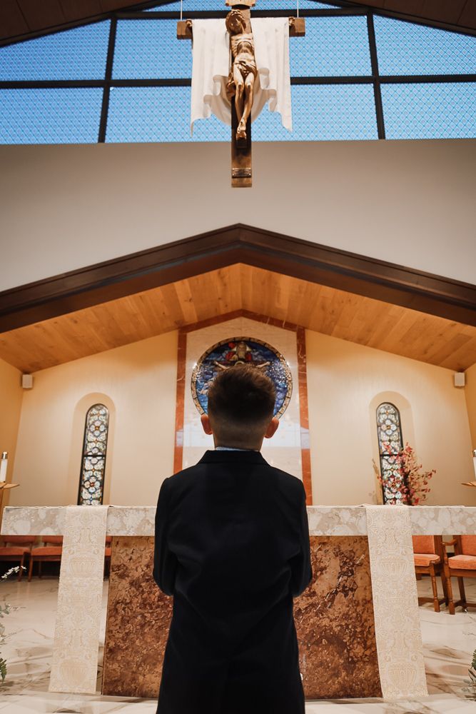Young boy in formal black suit standing before church altar with crucifix during First Communion ceremony.