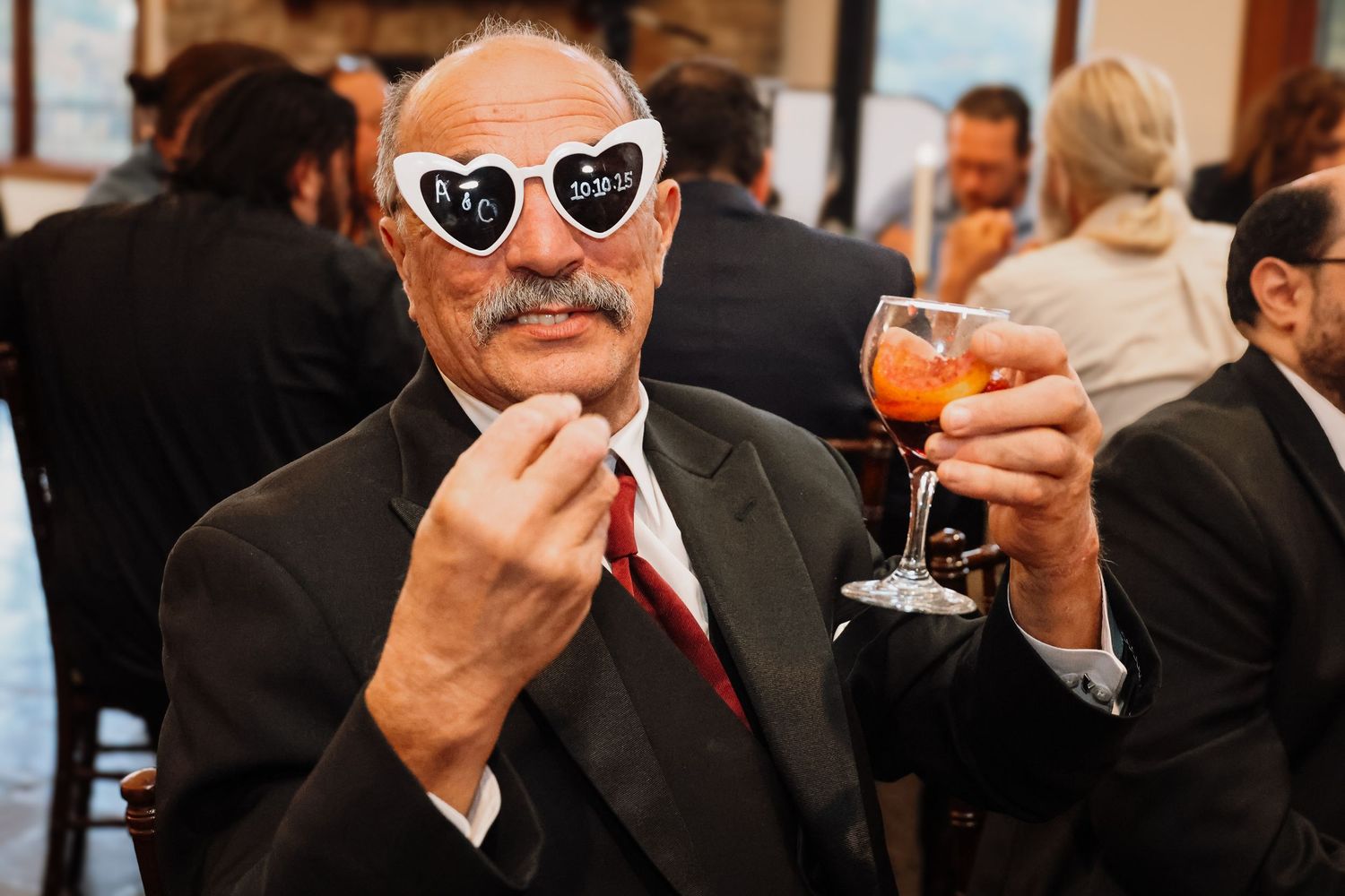 A person wearing heart-shaped sunglasses and a suit smiles while holding a cocktail at a social gathering.