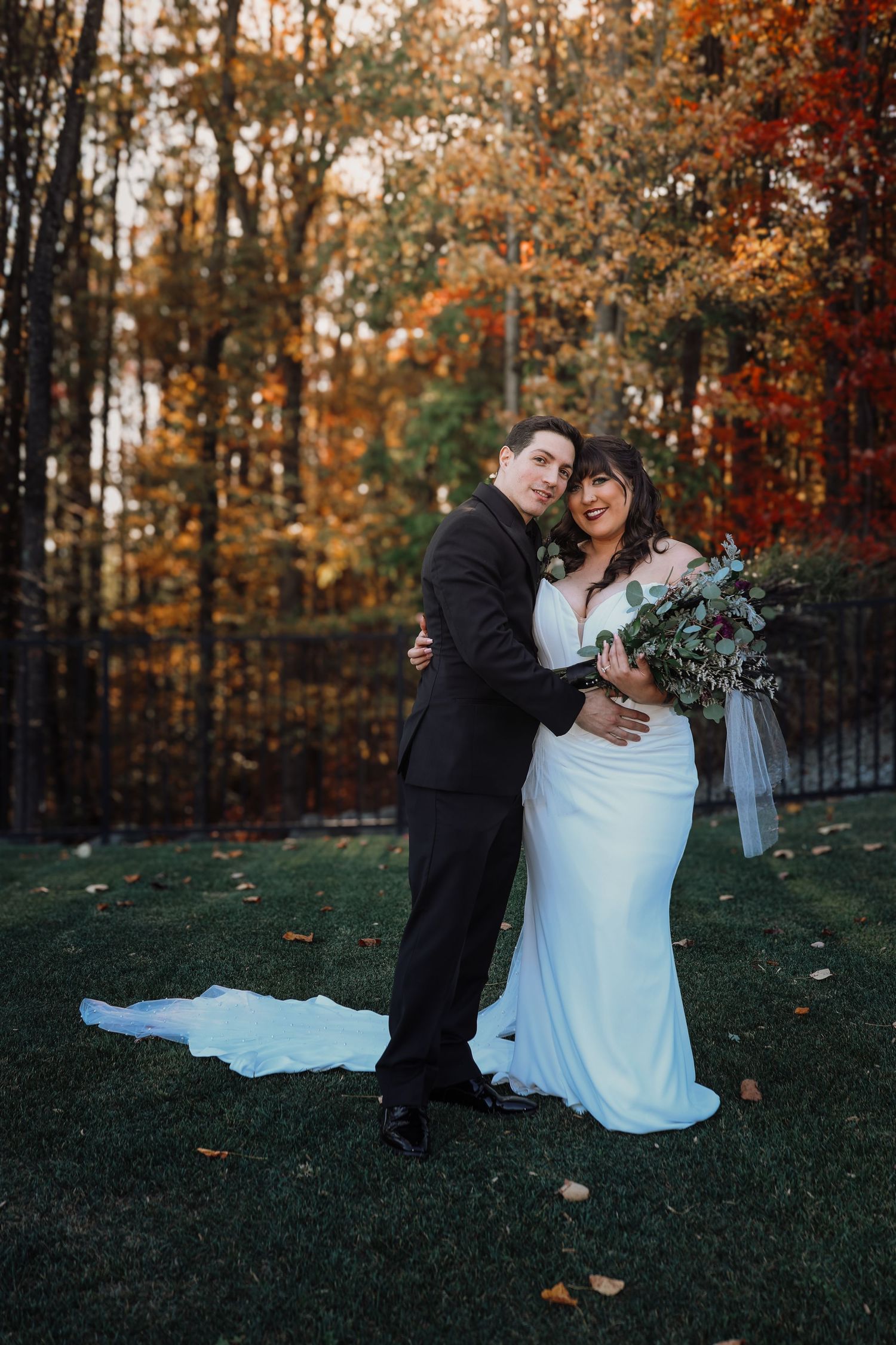 A couple in wedding attire shares intimate moments on grass surrounded by autumn trees with orange foliage.