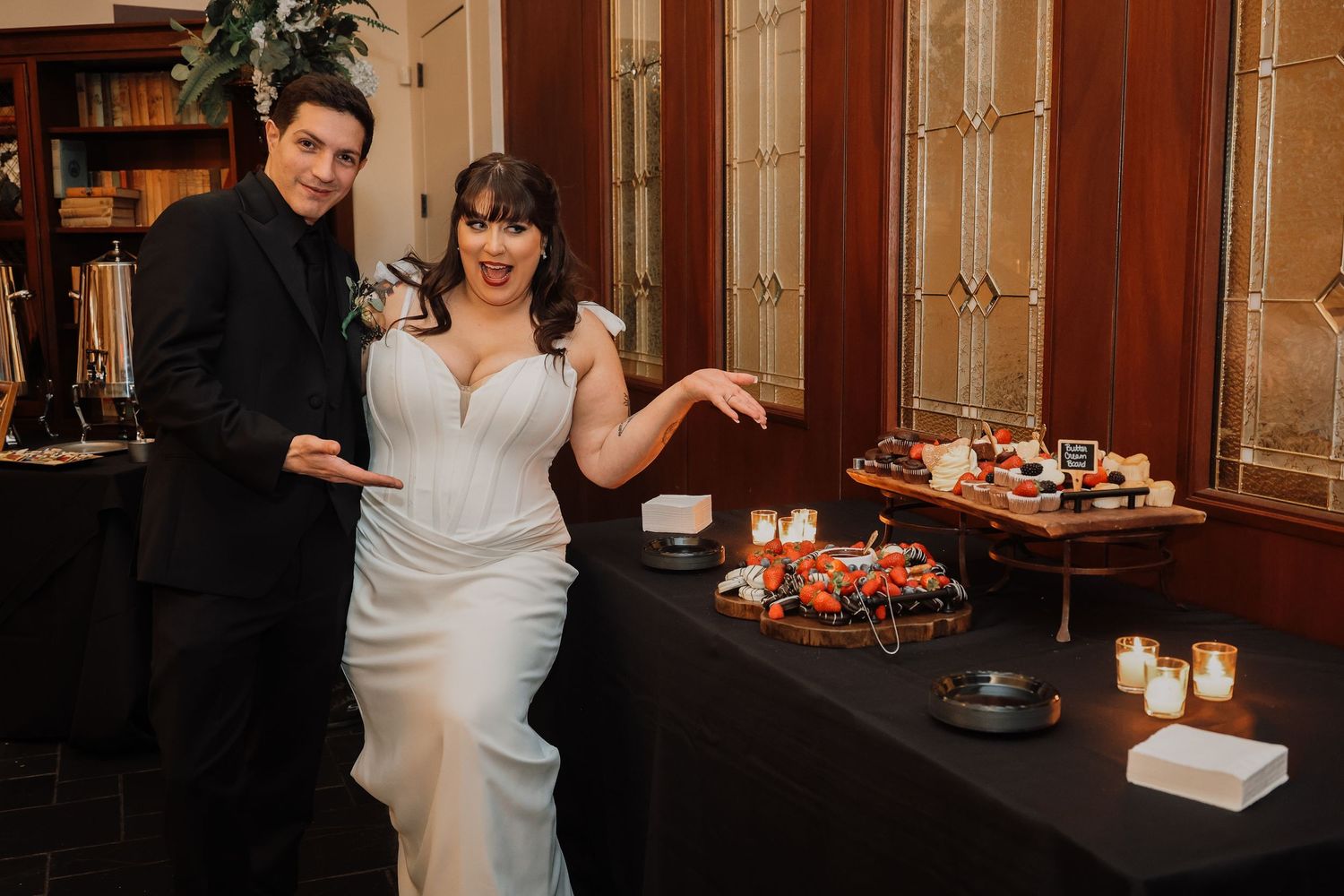 Couple in formal attire celebrates at wedding reception with dessert table and candles in elegant venue.