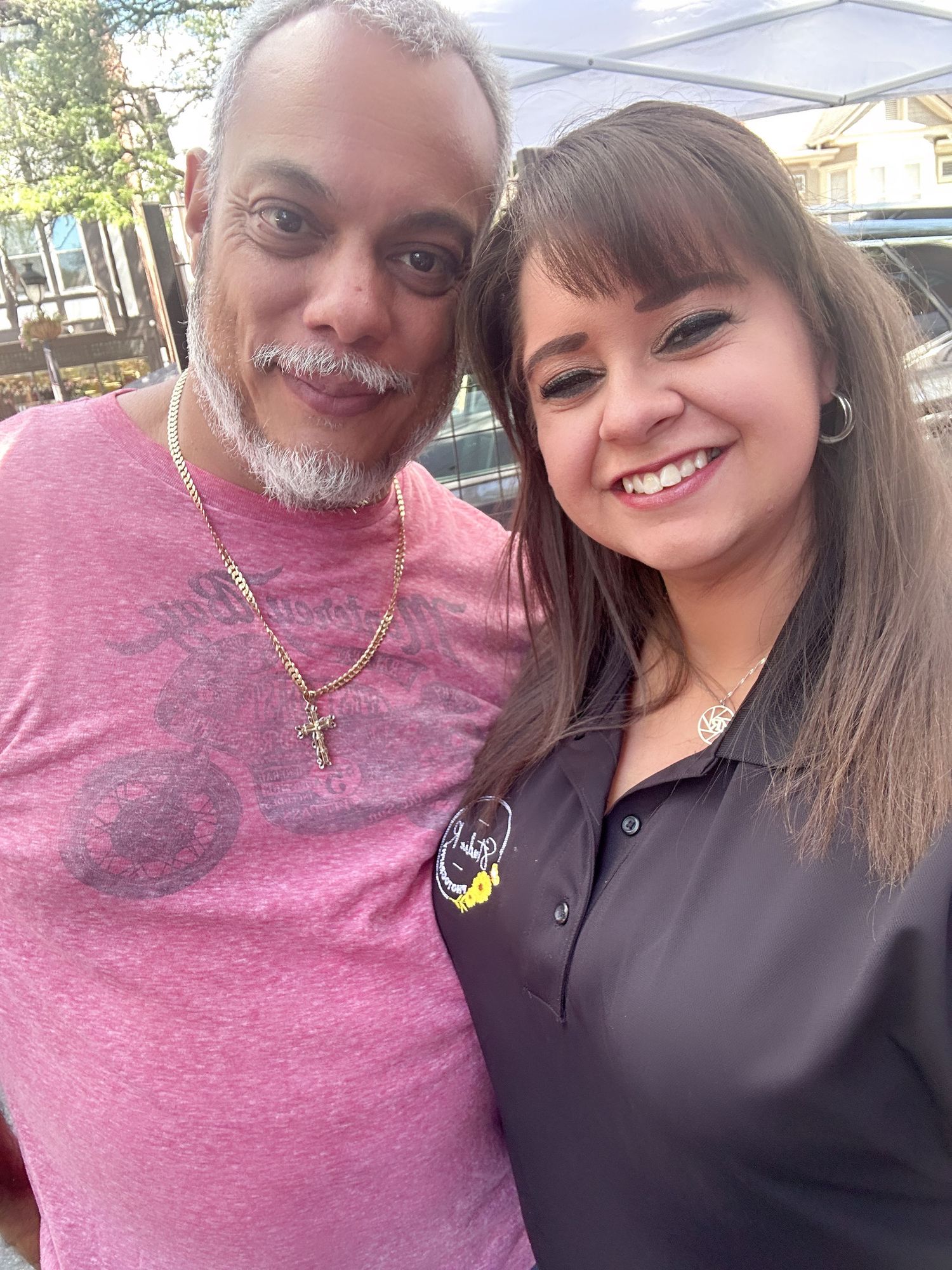 Two people sitting close together outside and smiling at the camera in casual attire.