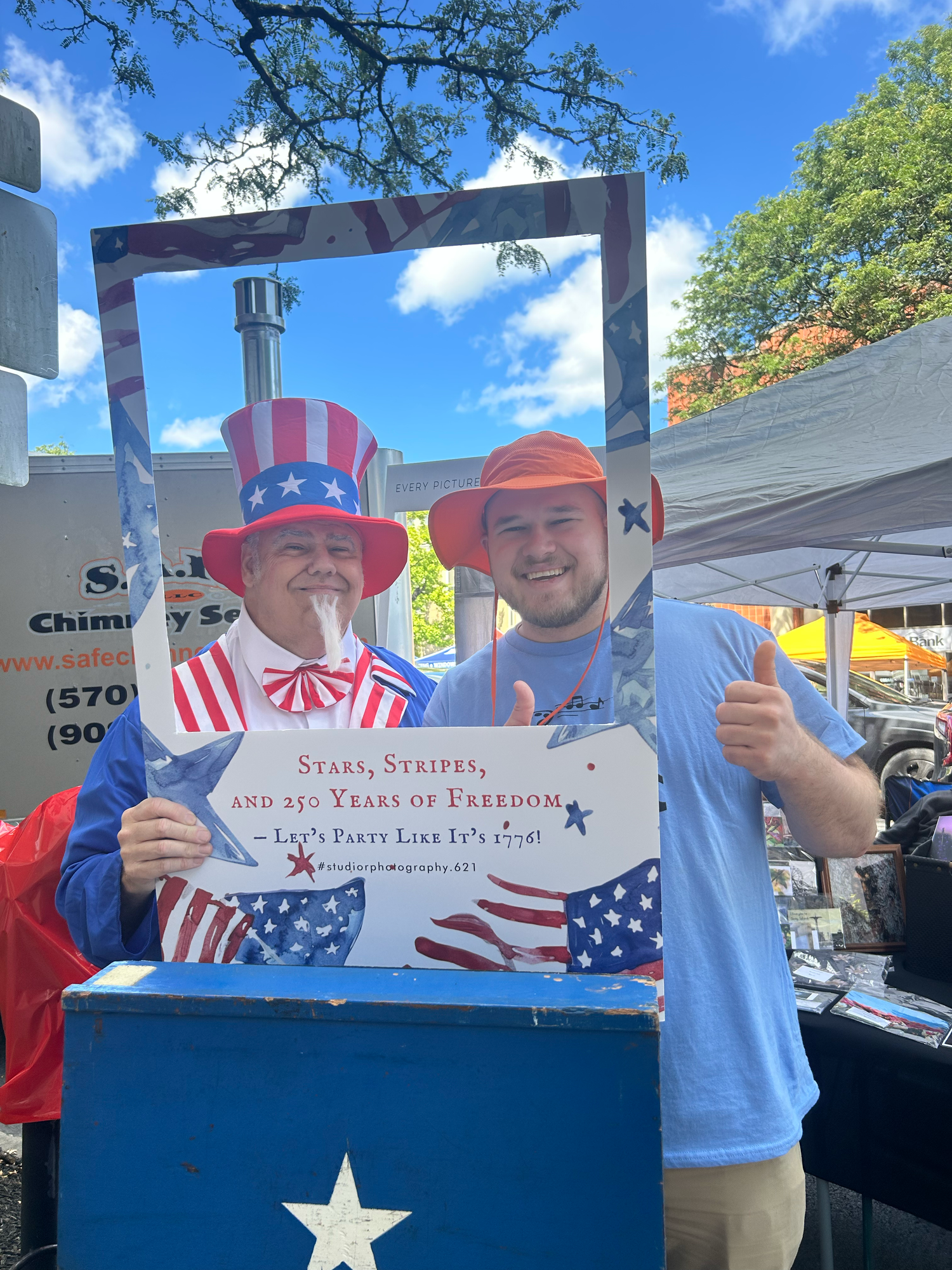 Group celebrating at an outdoor patriotic event with American flag themed clothing and signs on a sunny day.