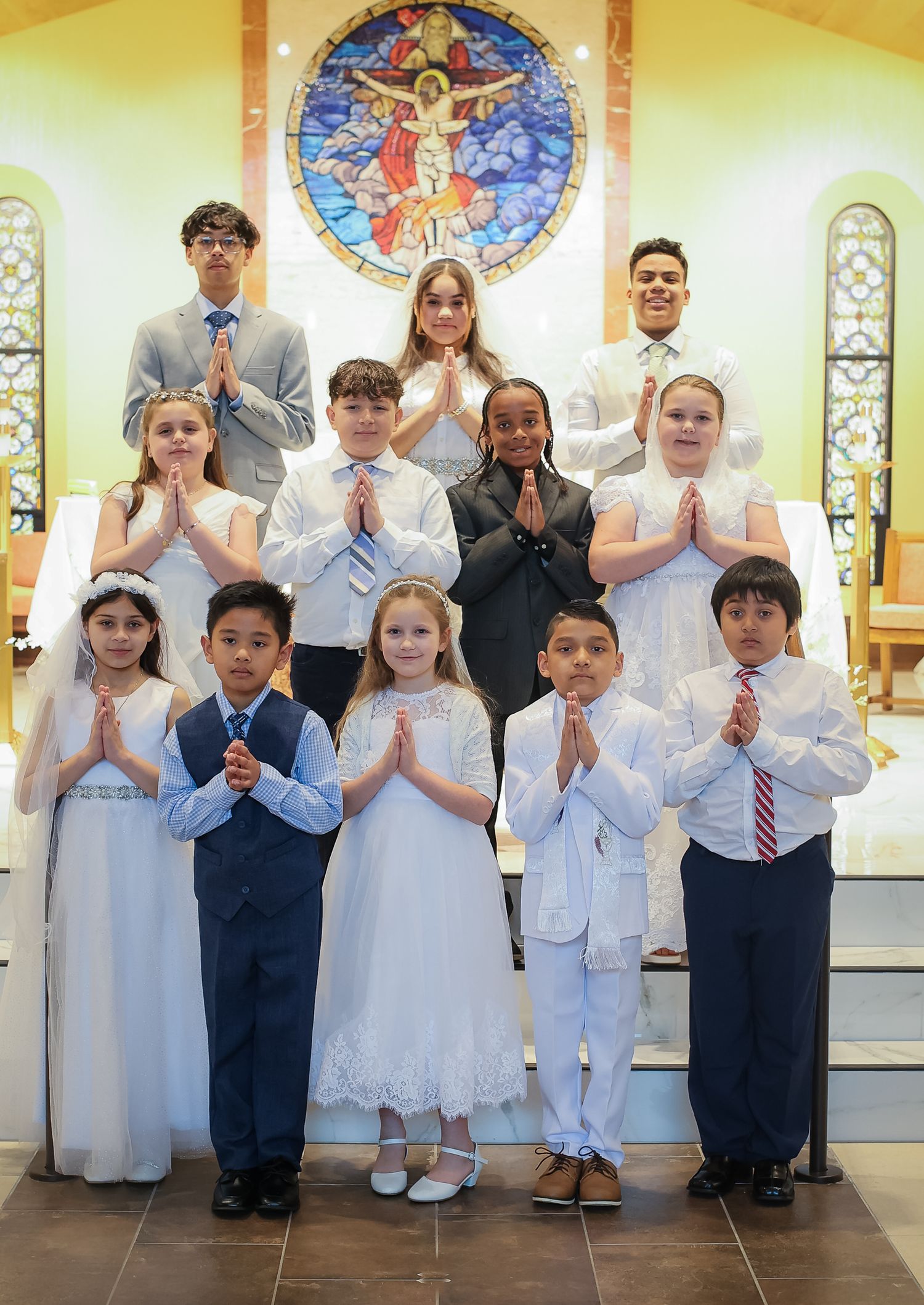 Children in white dresses and suits stand in a line at church for their First Communion ceremony.