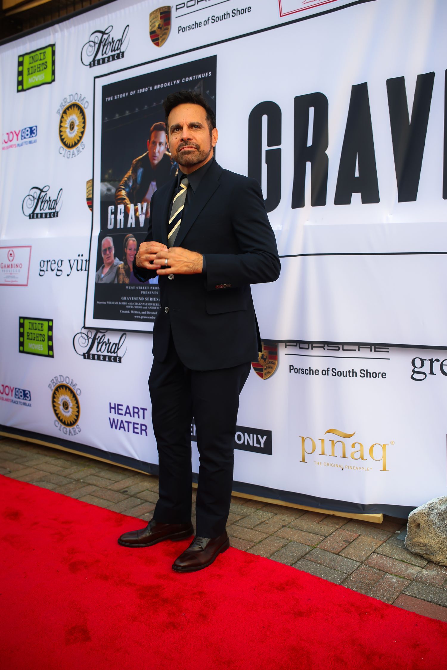 Person in black suit stands on red carpet at event with sponsor logos and step-and-repeat backdrop.