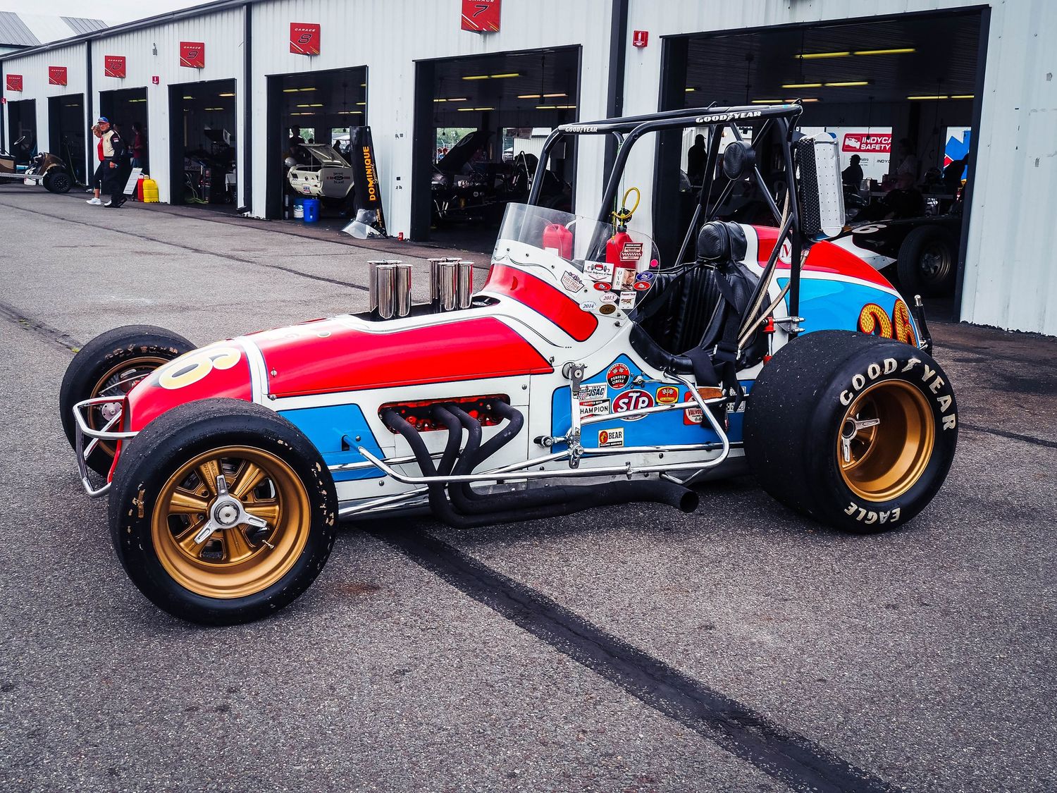 A vintage red, white and blue sprint car with gold wheels sits parked in front of racing garage bays.