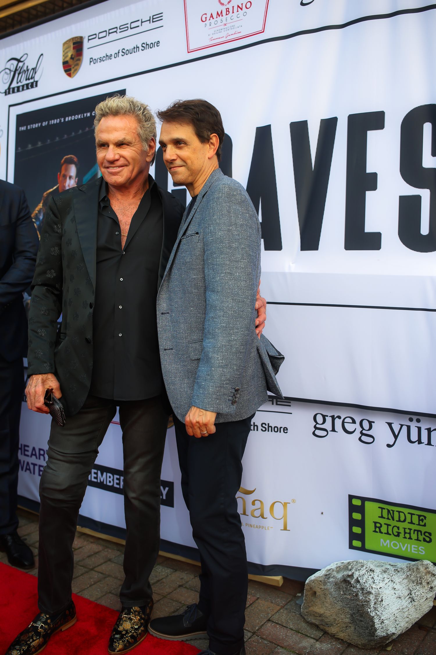 Two people in dark jackets pose together at a red carpet event with sponsor logos visible on the backdrop.