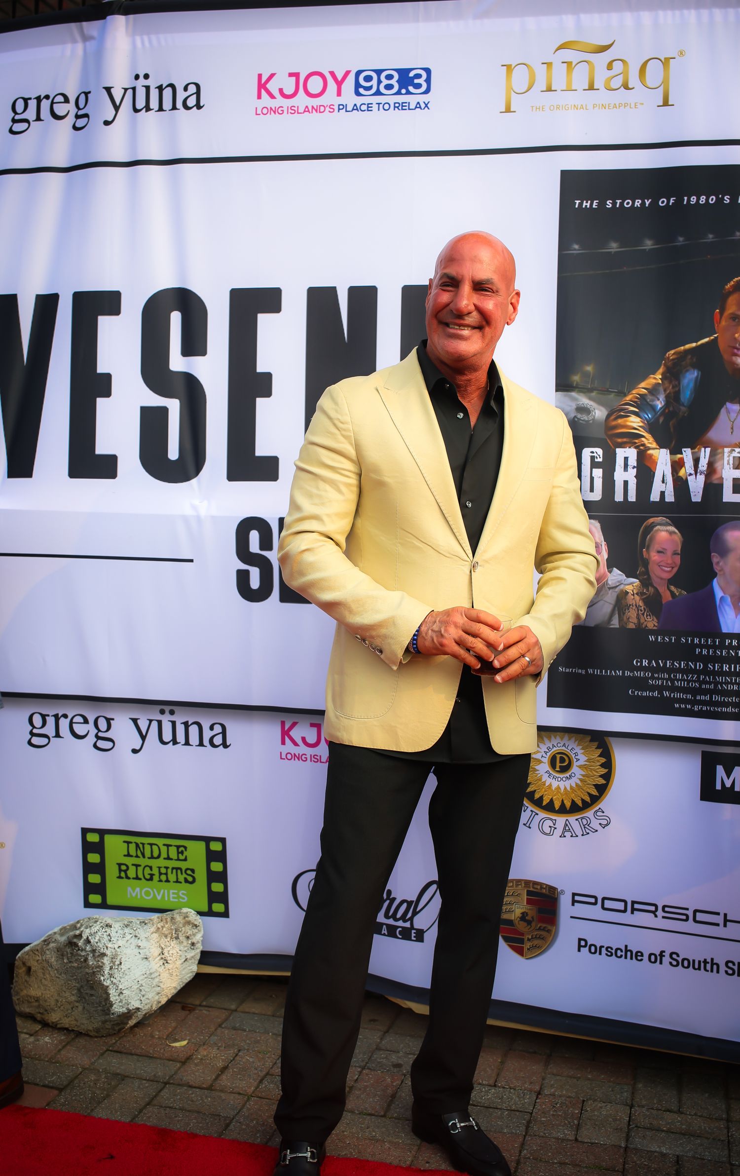 A person in a yellow blazer and dark pants stands on a red carpet at an event with promotional banners and signage.