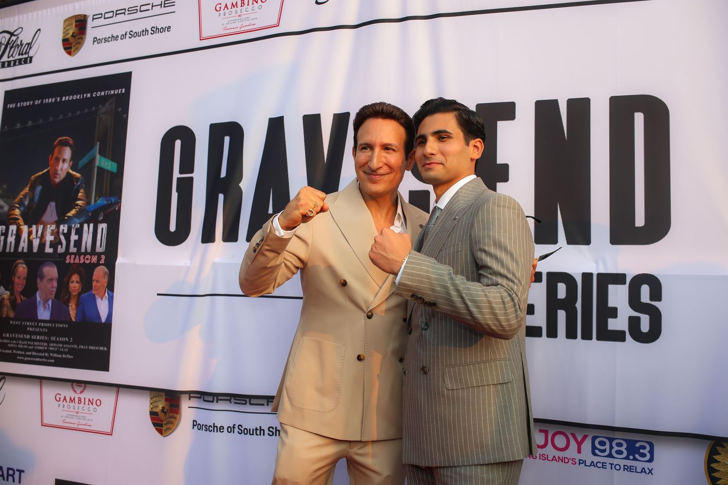 Two people in suits pose together at a premiere event in front of a backdrop with text reading GRAND.