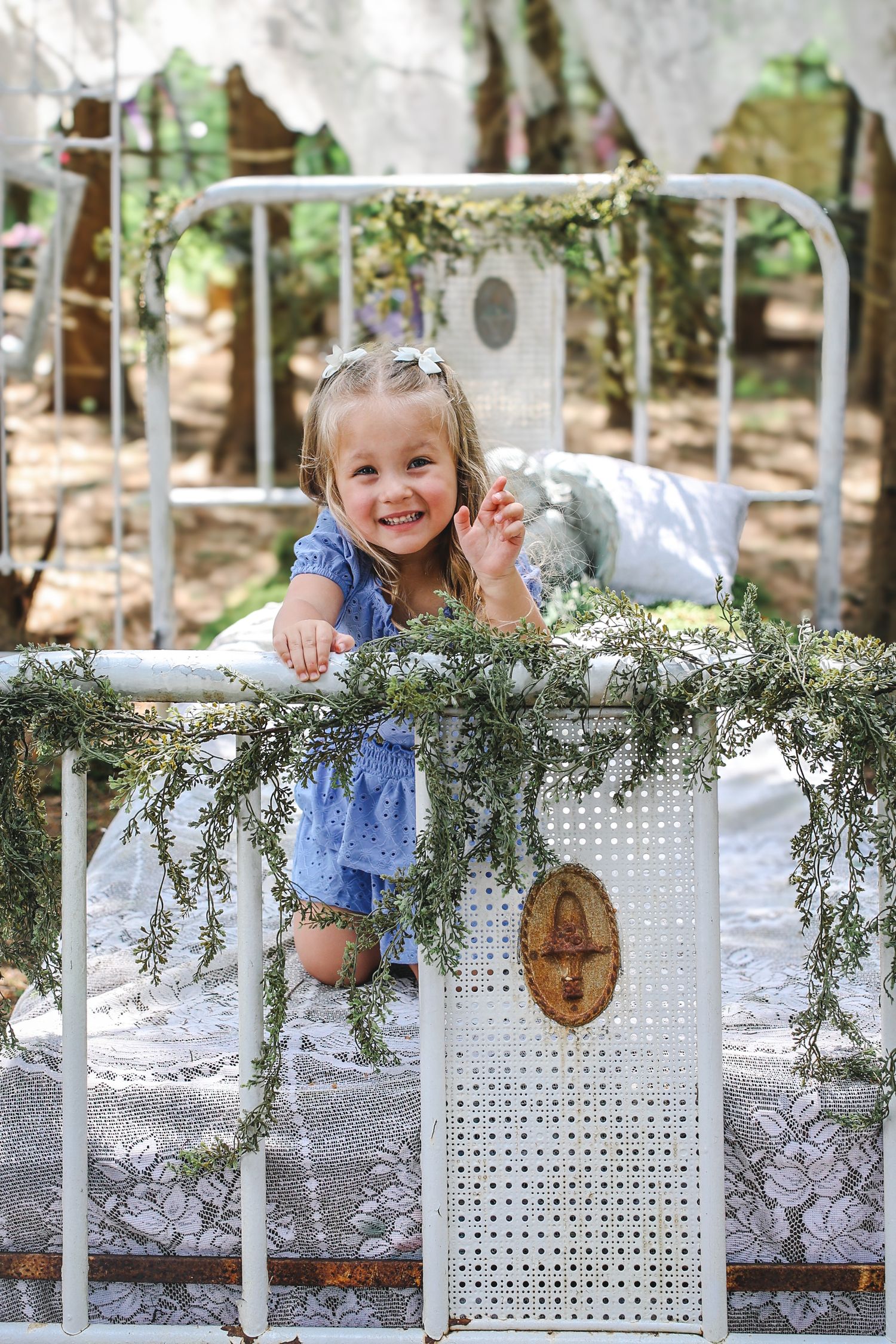 A child in a blue dress peeks playfully through decorative greenery draped across white outdoor railings.